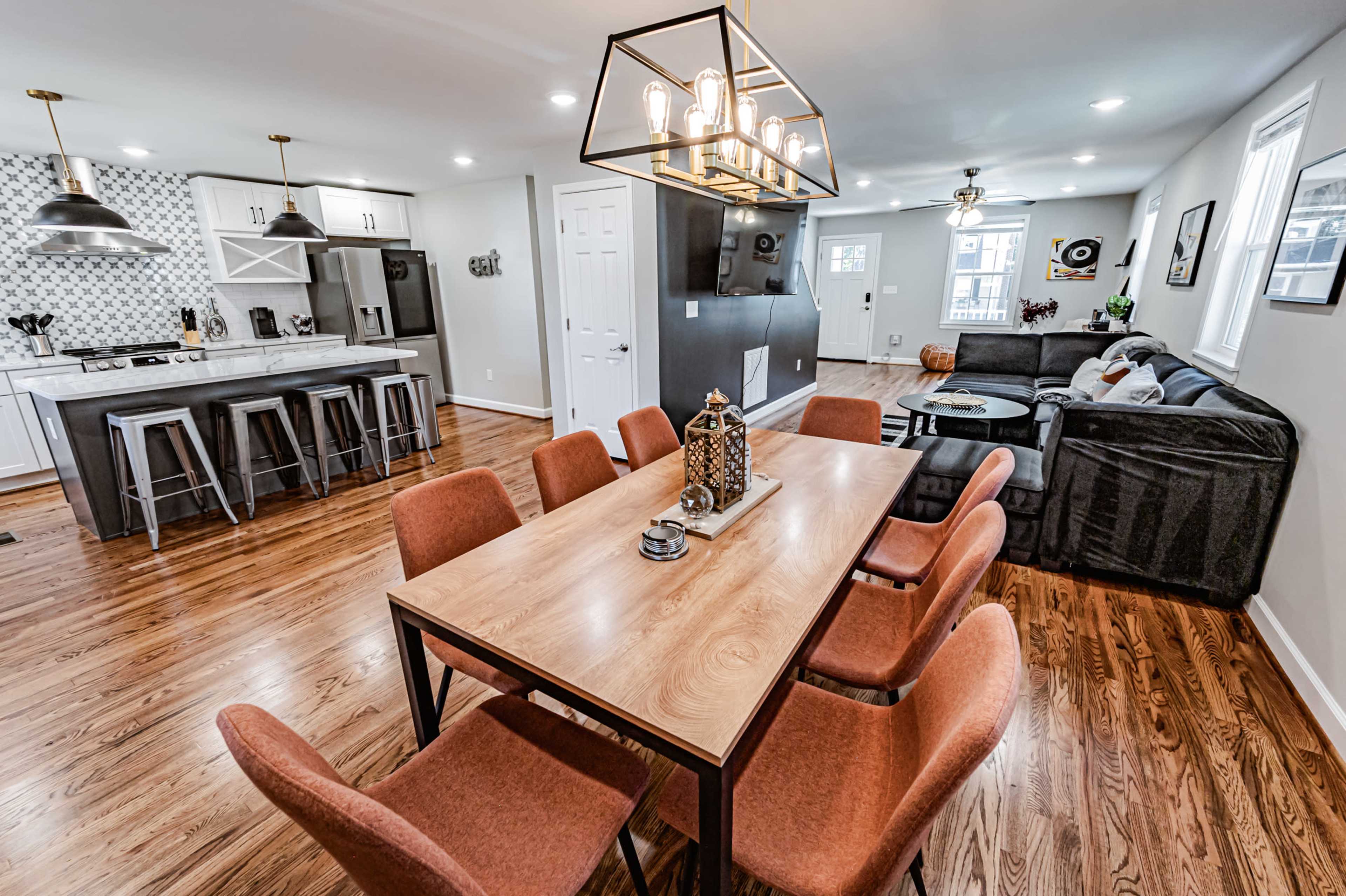 A modern open-concept living space featuring a dining area with a wooden table and eight orange chairs, adjacent to a kitchen with white cabinets and stainless steel appliances.
