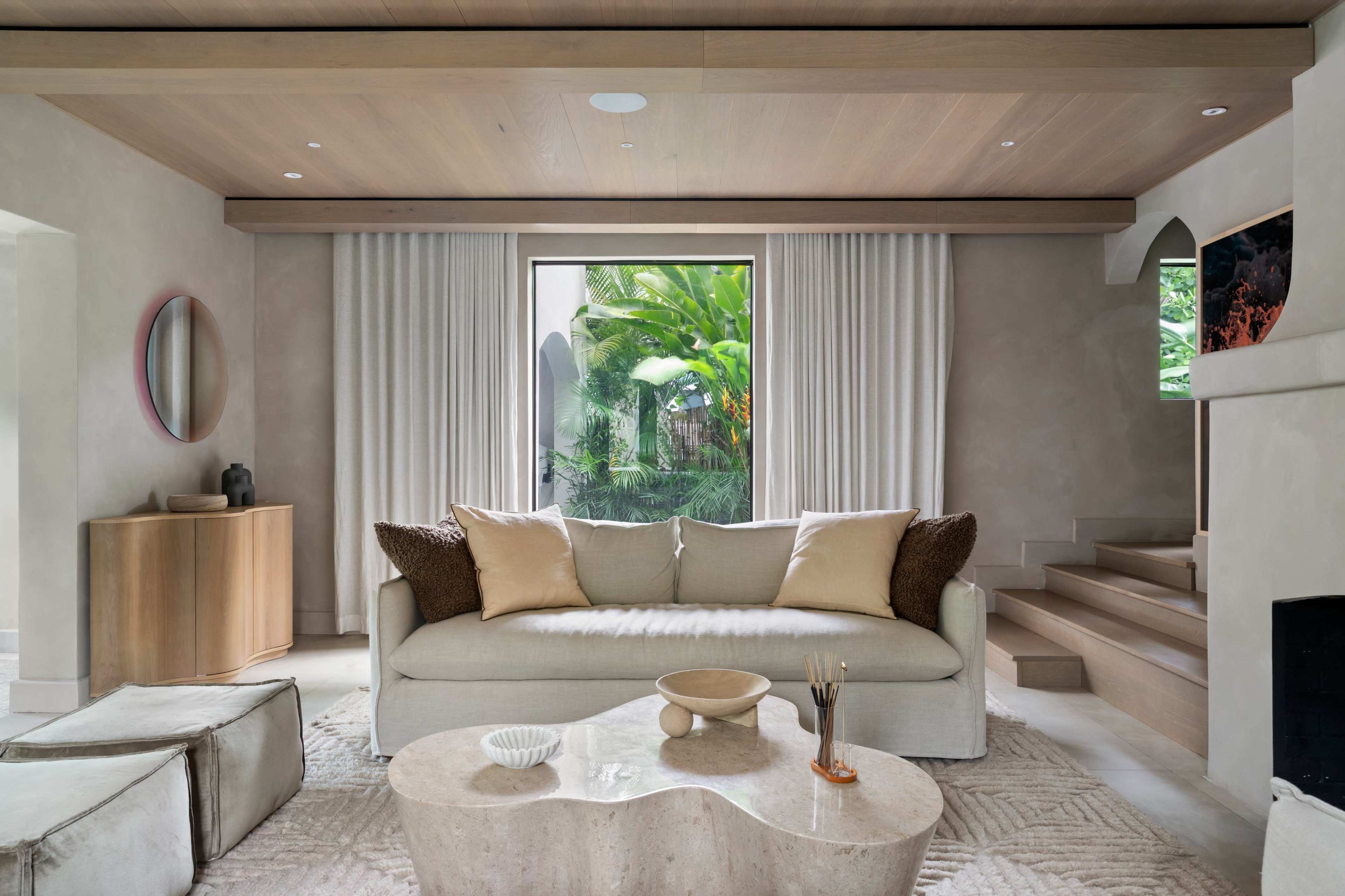 A modern living room features a light-colored sofa with decorative pillows, a round coffee table, and large windows with greenery visible outside.