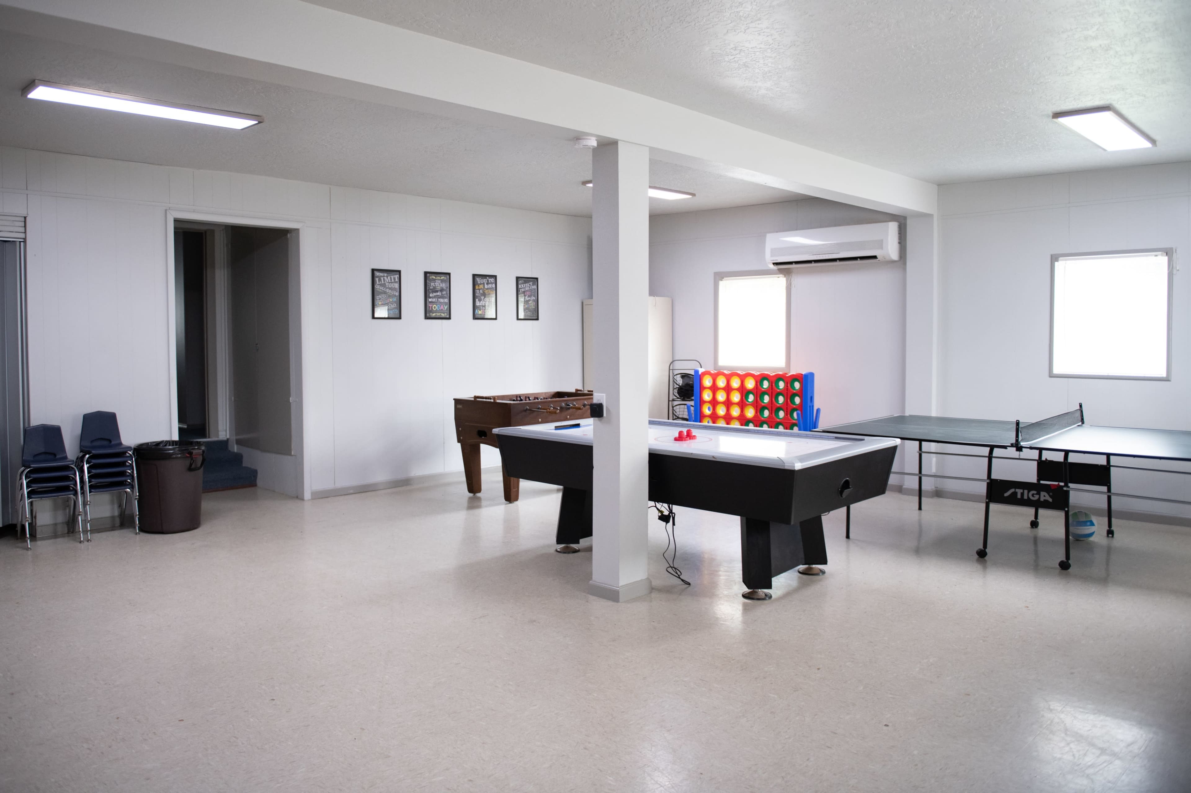 A recreation room featuring a foosball table, a connect four game, and a ping pong table in a well-lit space with beige walls and a door leading to another area.