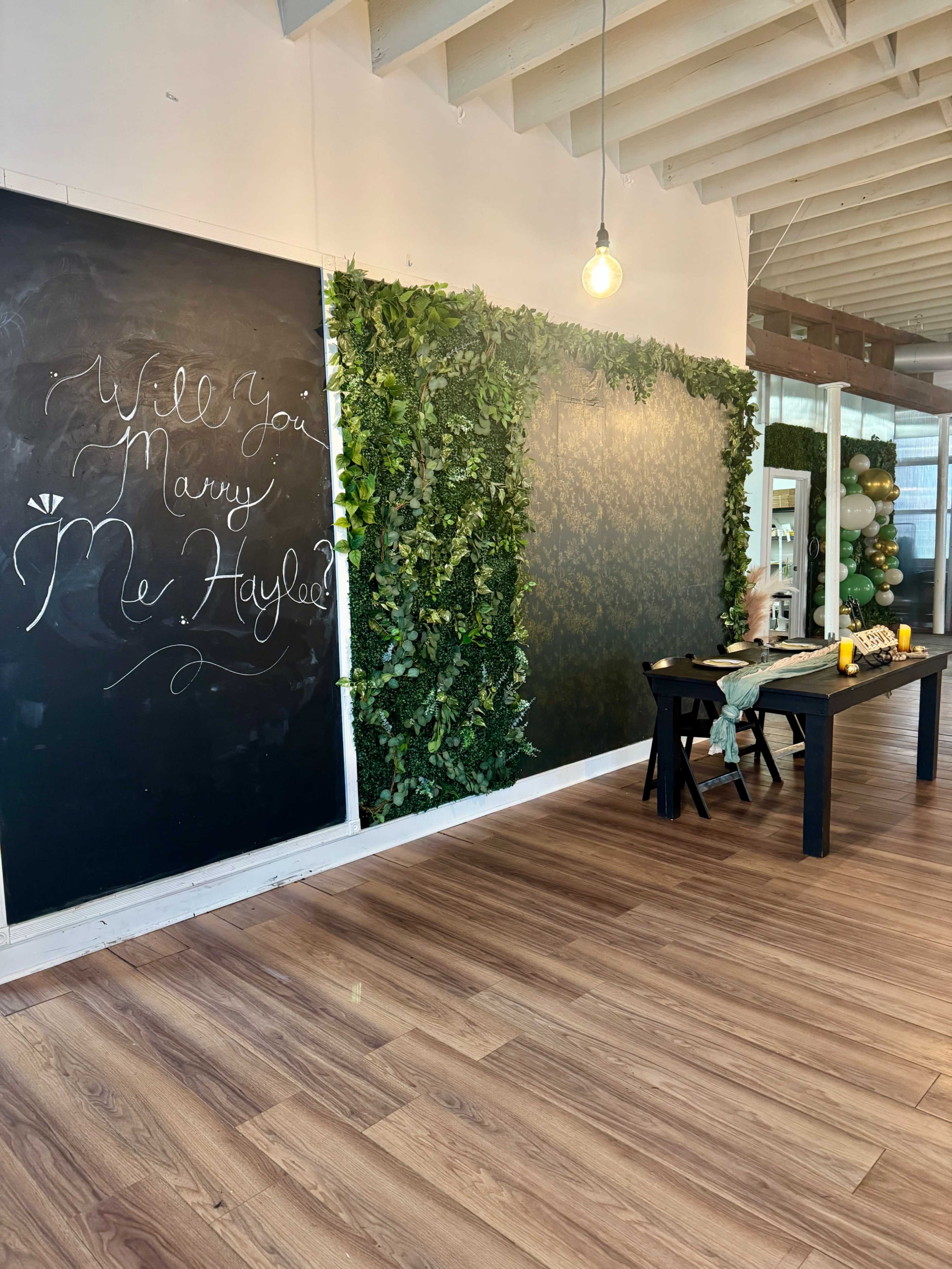 The image shows a room with a chalkboard wall displaying the text "Will You Marry Me Hayley," greenery along one side, and a table set with decorative elements.