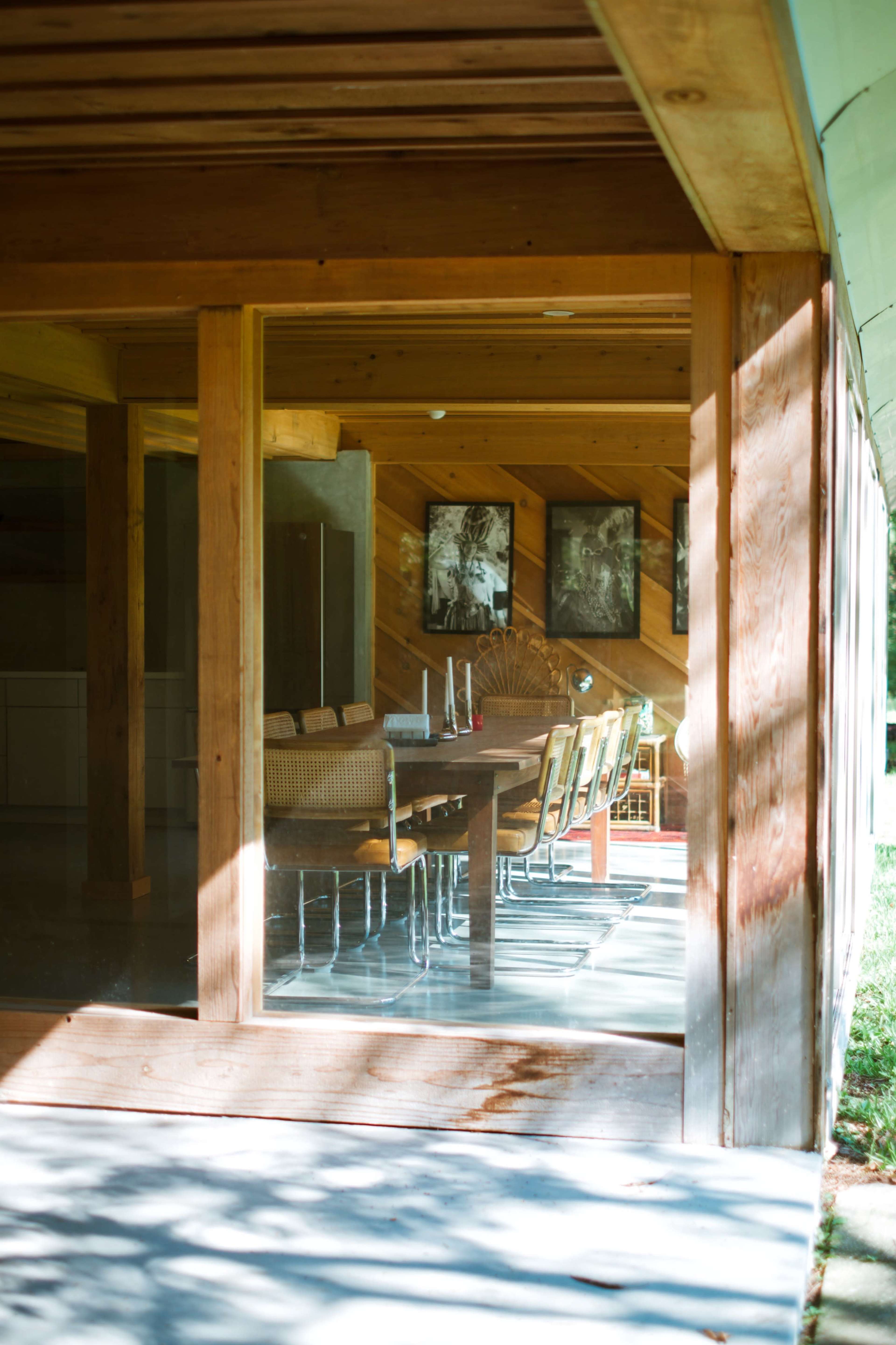 A wooden dining area with a long table and several chairs is visible through a large glass window, framed by natural wood siding.