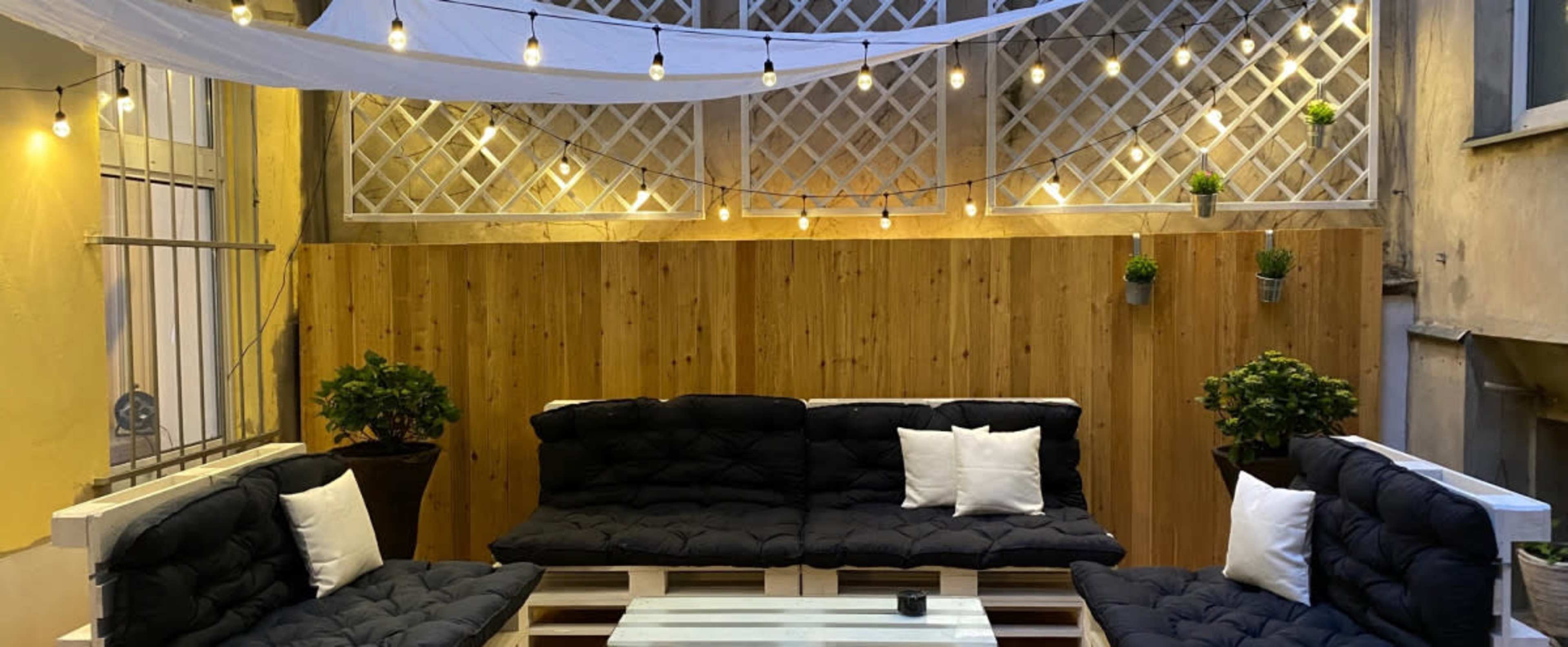 A cozy outdoor seating area with dark cushioned sofas arranged around a white coffee table, illuminated by string lights and draped fabric overhead.