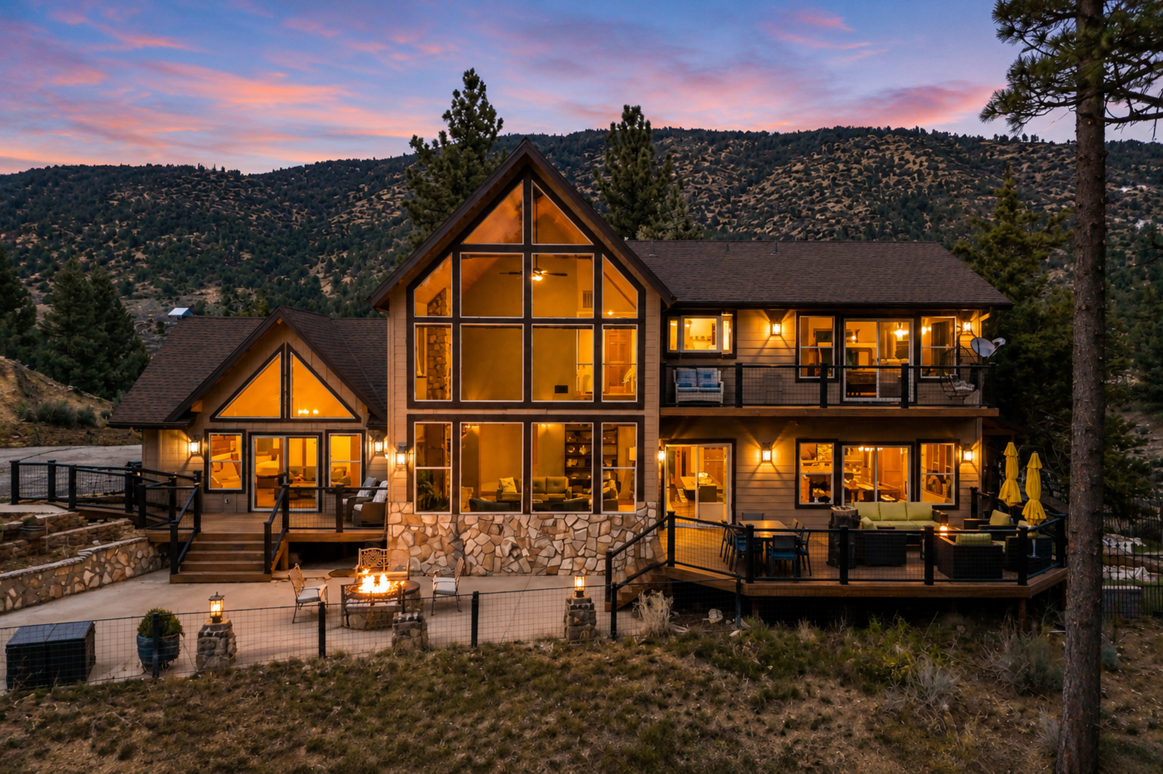 A modern chalet-style house with large windows sits against a backdrop of mountains at sunset, featuring a patio with seating and a stone fire pit.