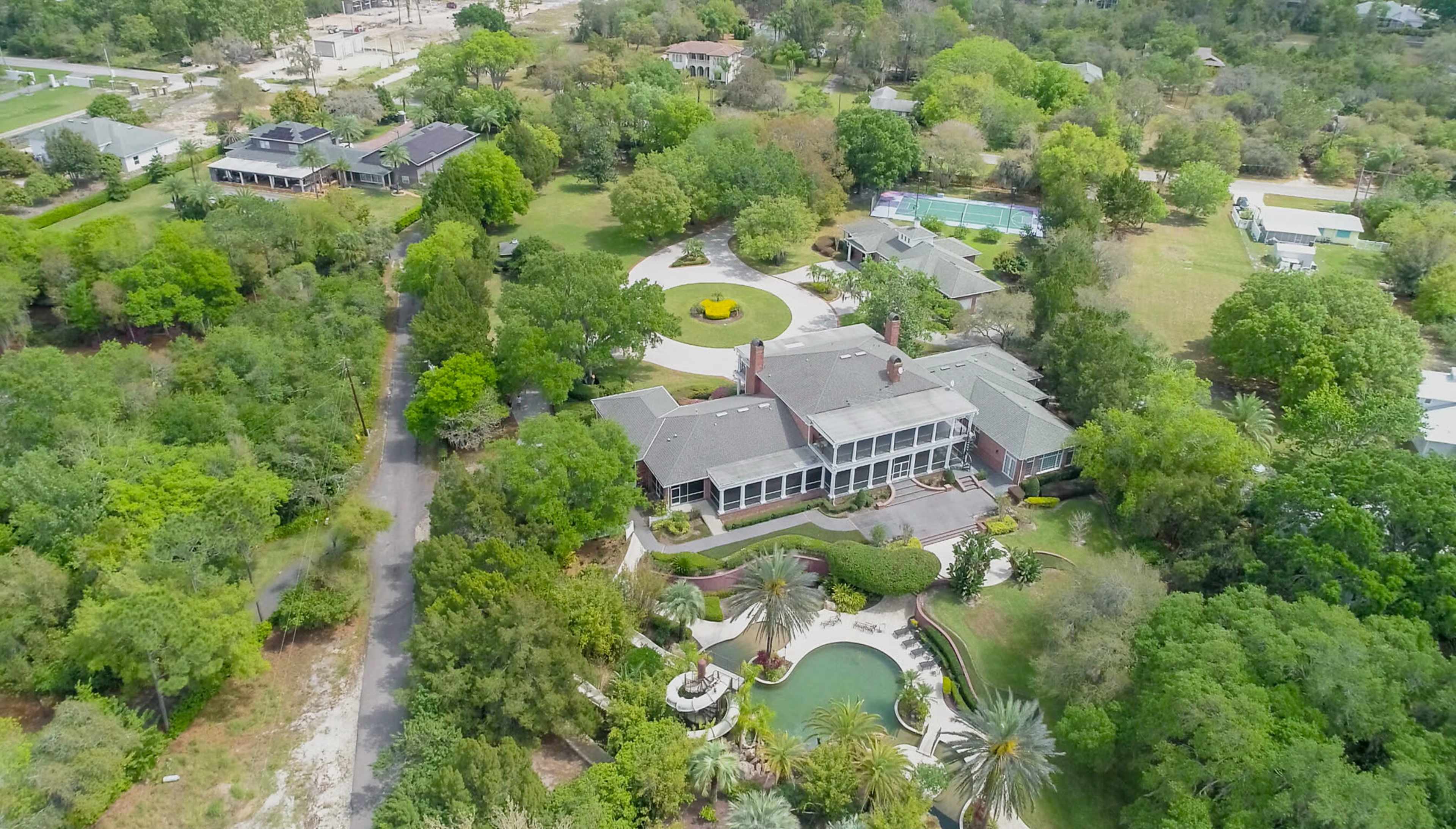 A large, sprawling estate surrounded by lush greenery and featuring a circular driveway, a pool, and landscaped gardens.