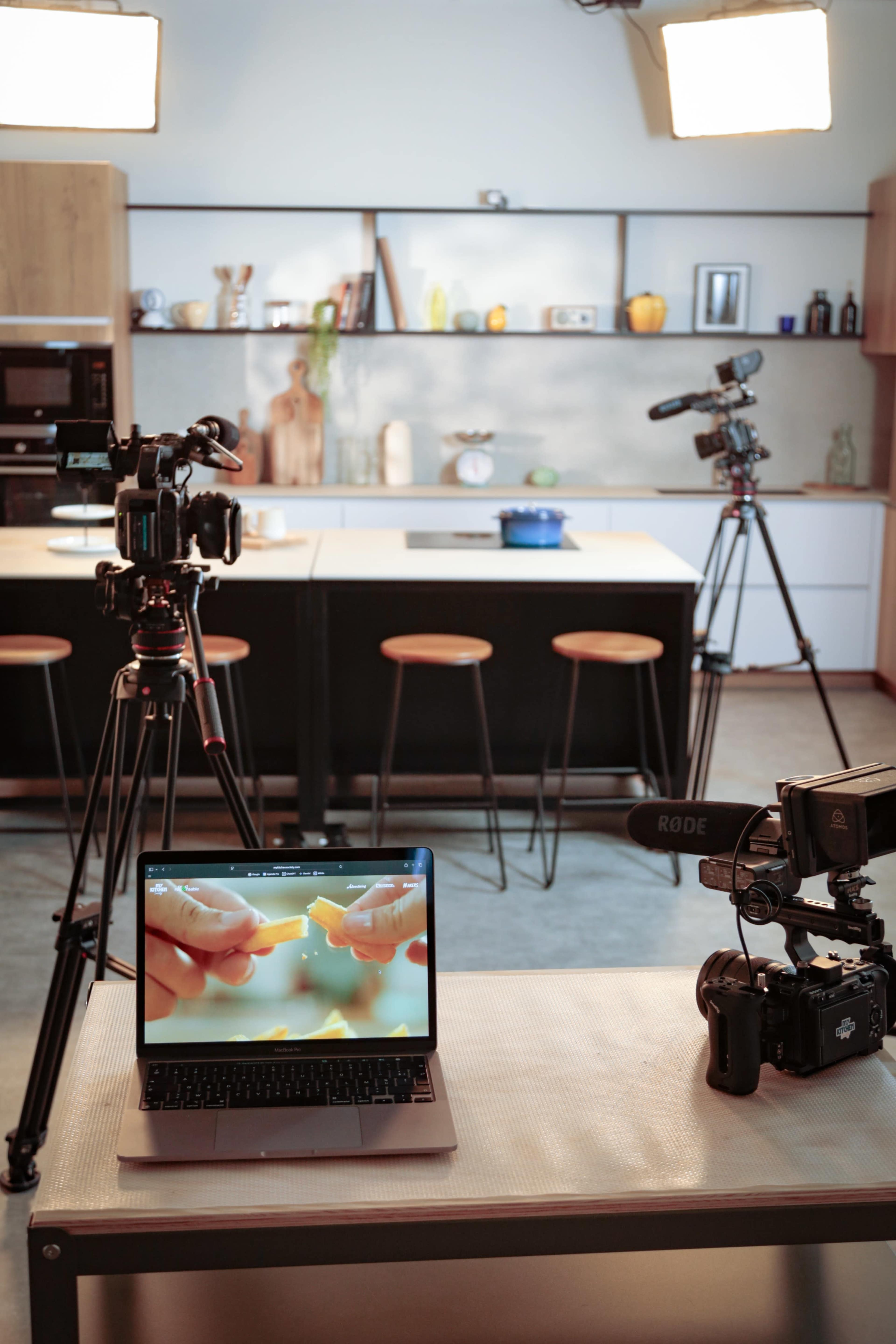 The image shows a modern kitchen setup with multiple cameras on tripods, a laptop displaying hands preparing food, and bright lights overhead.