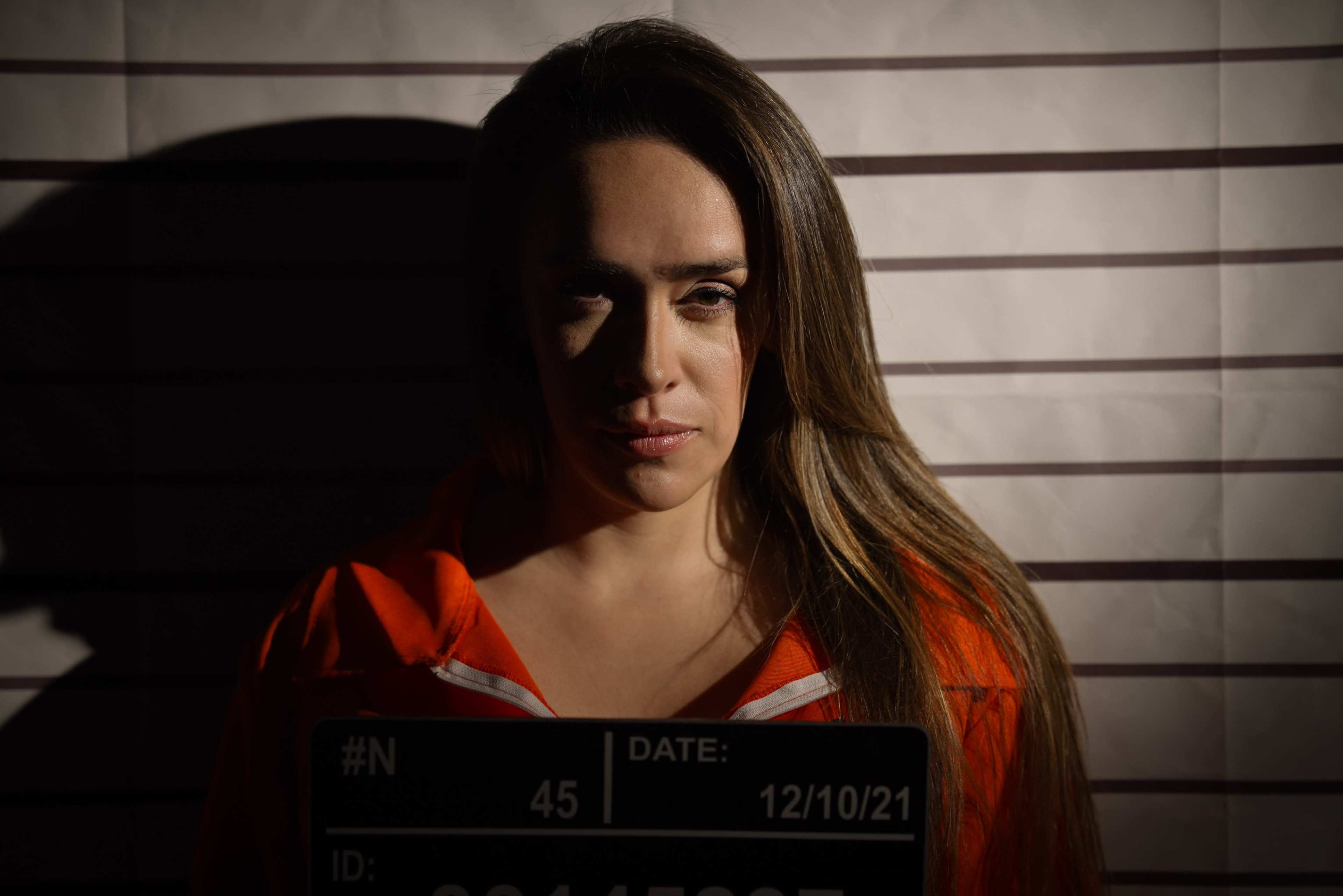 A woman wearing an orange jumpsuit stands against a striped backdrop, holding a mugshot board with her details.