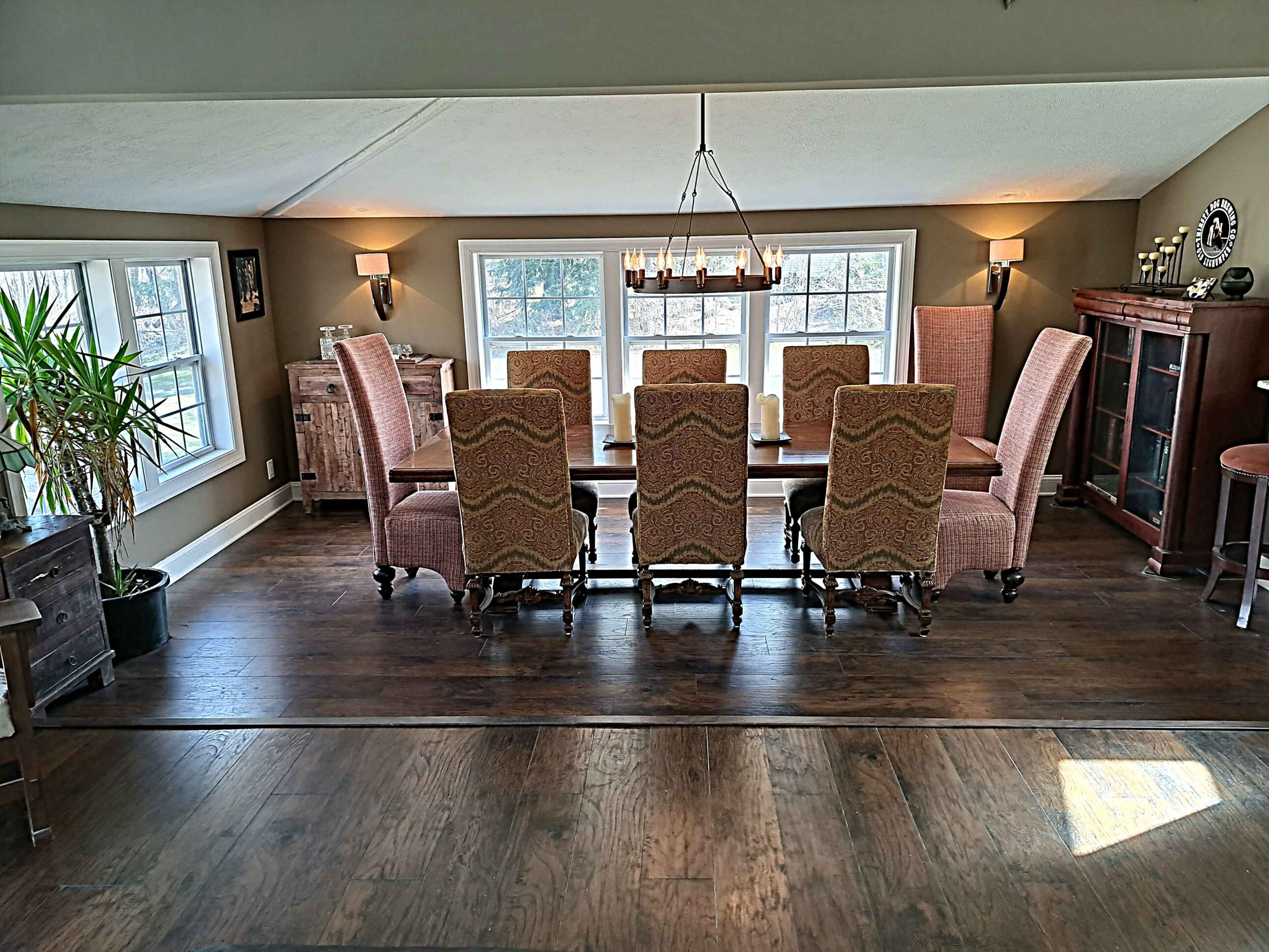 A large dining room features a long wooden table surrounded by eight patterned chairs, with a window providing natural light and a plant in the corner.