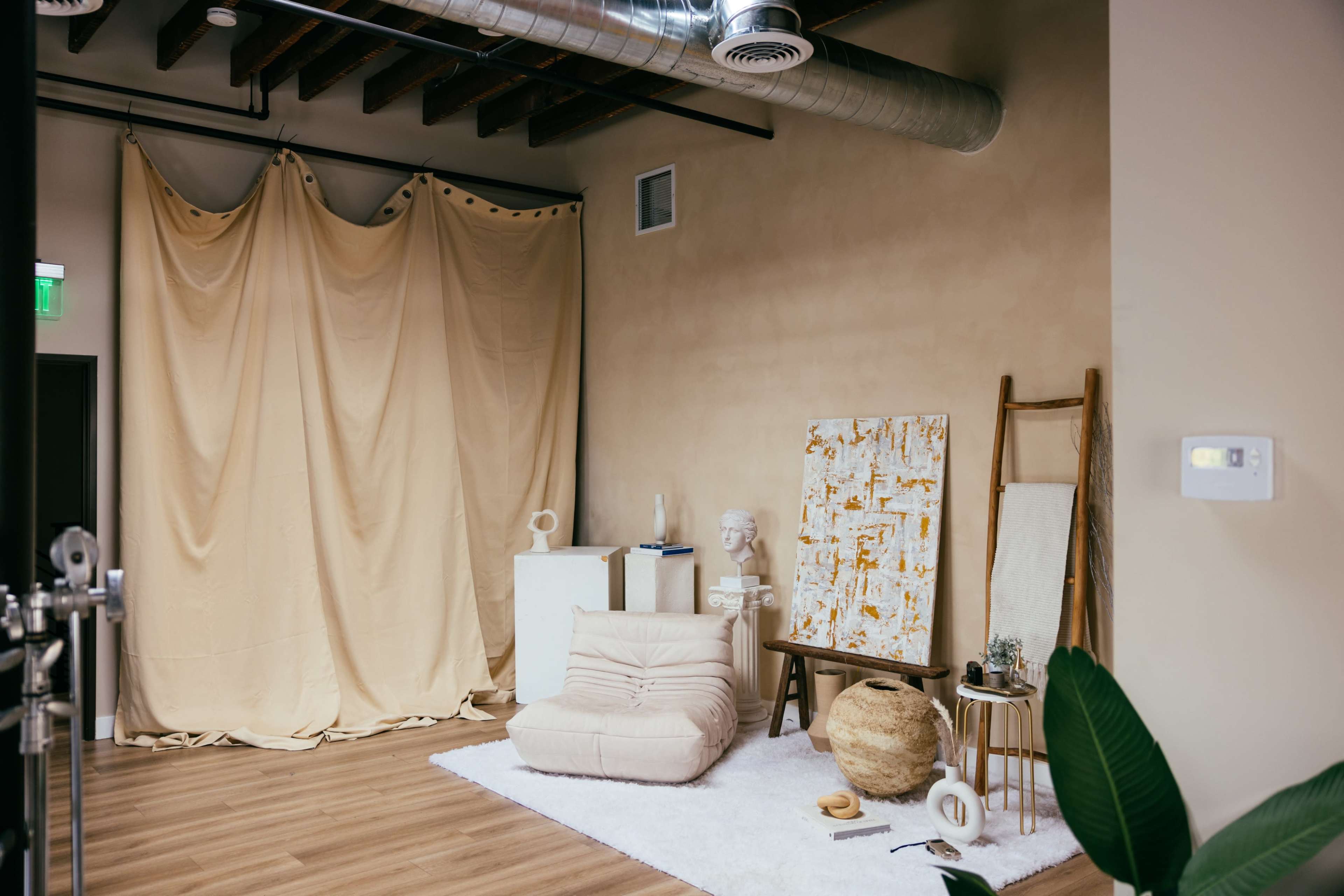 The image shows a cozy interior space with beige walls, a large curtain draped against the back, a cream-colored lounge chair, a textured rug, and various art pieces and decor items.