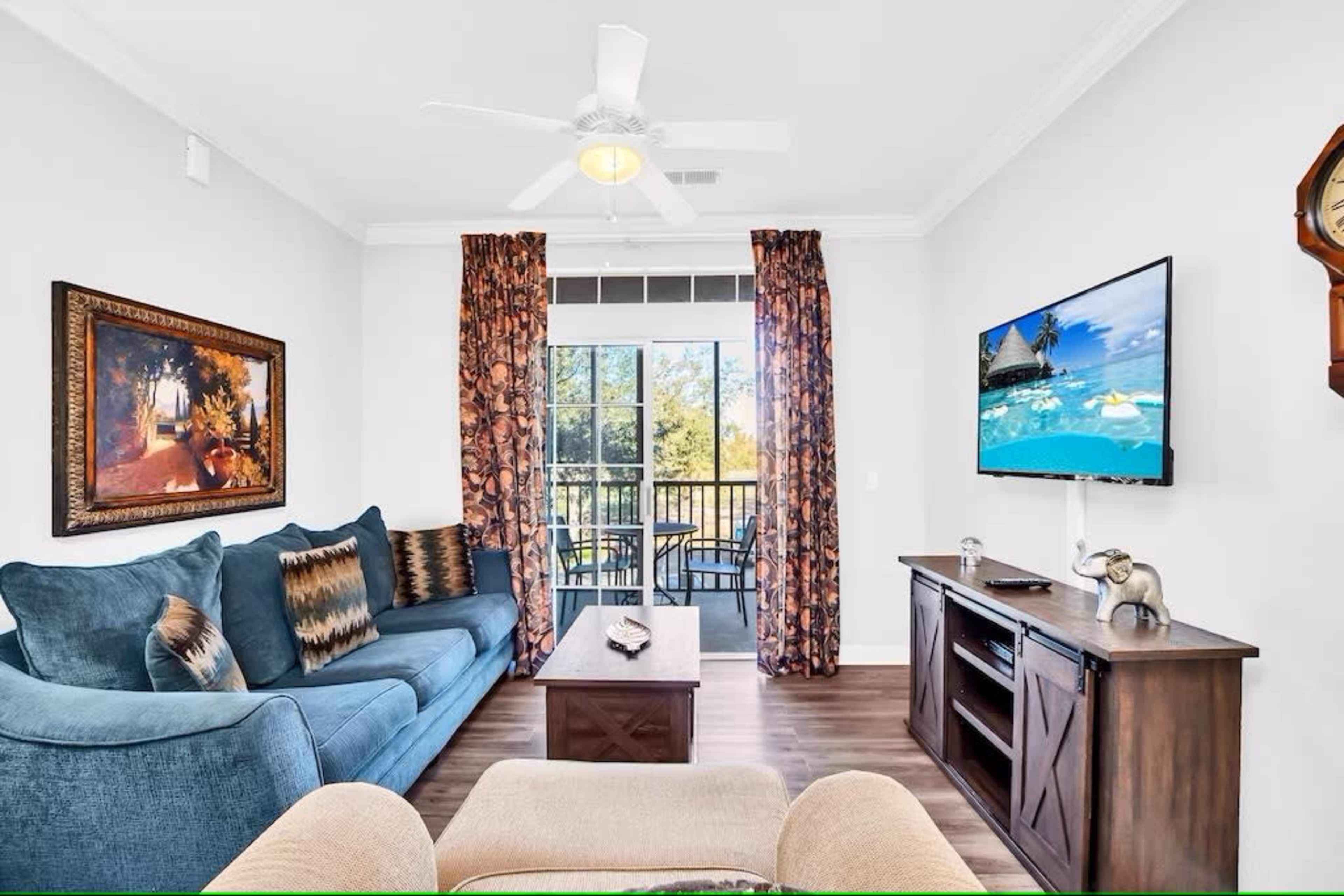 A living room features a blue sofa, a wooden coffee table, a television mounted on the wall, and curtains with floral patterns, overlooking a balcony through large windows.