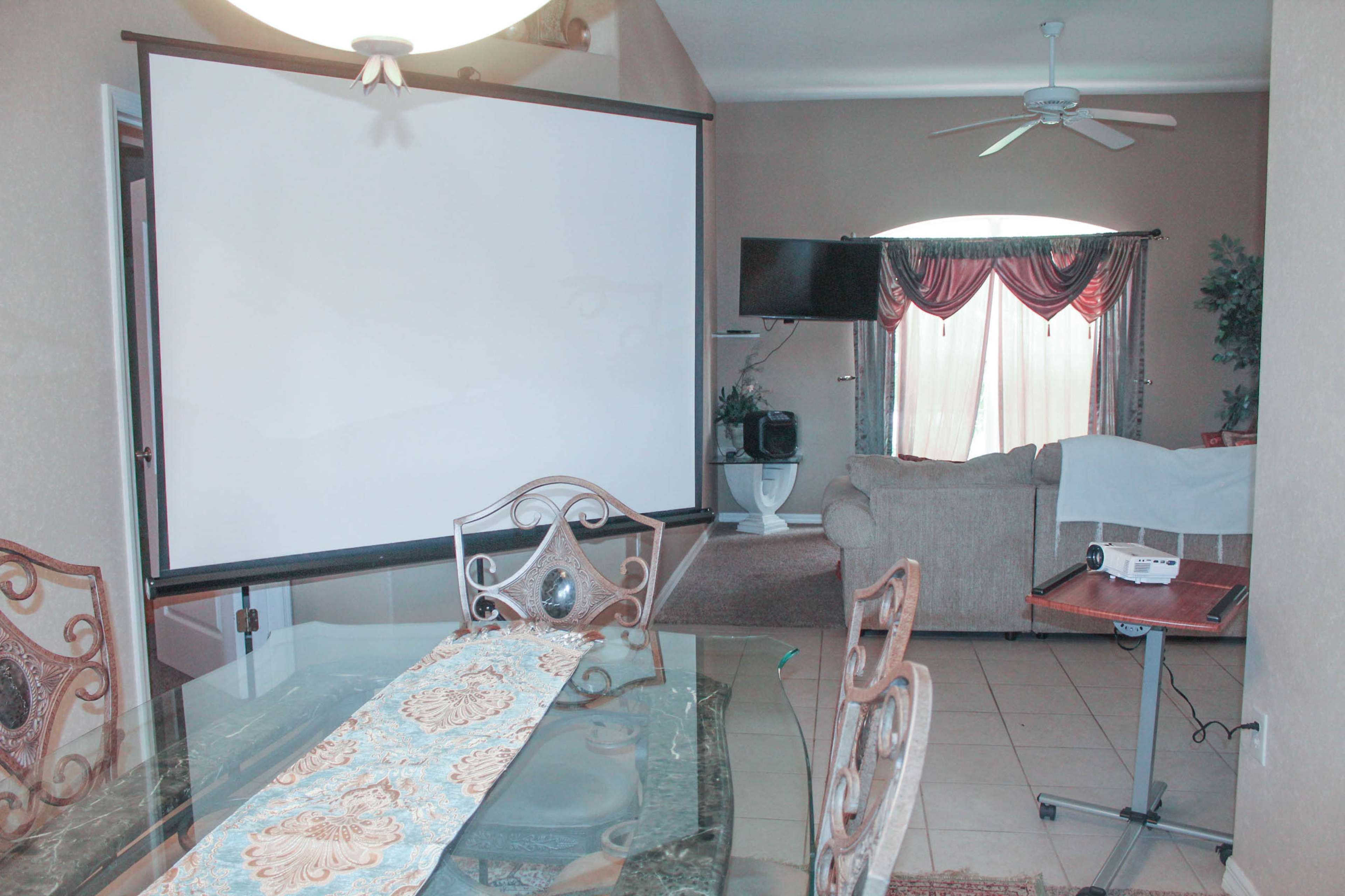 A dining area with a glass table and chairs adjacent to a living room featuring a projector screen and a TV.