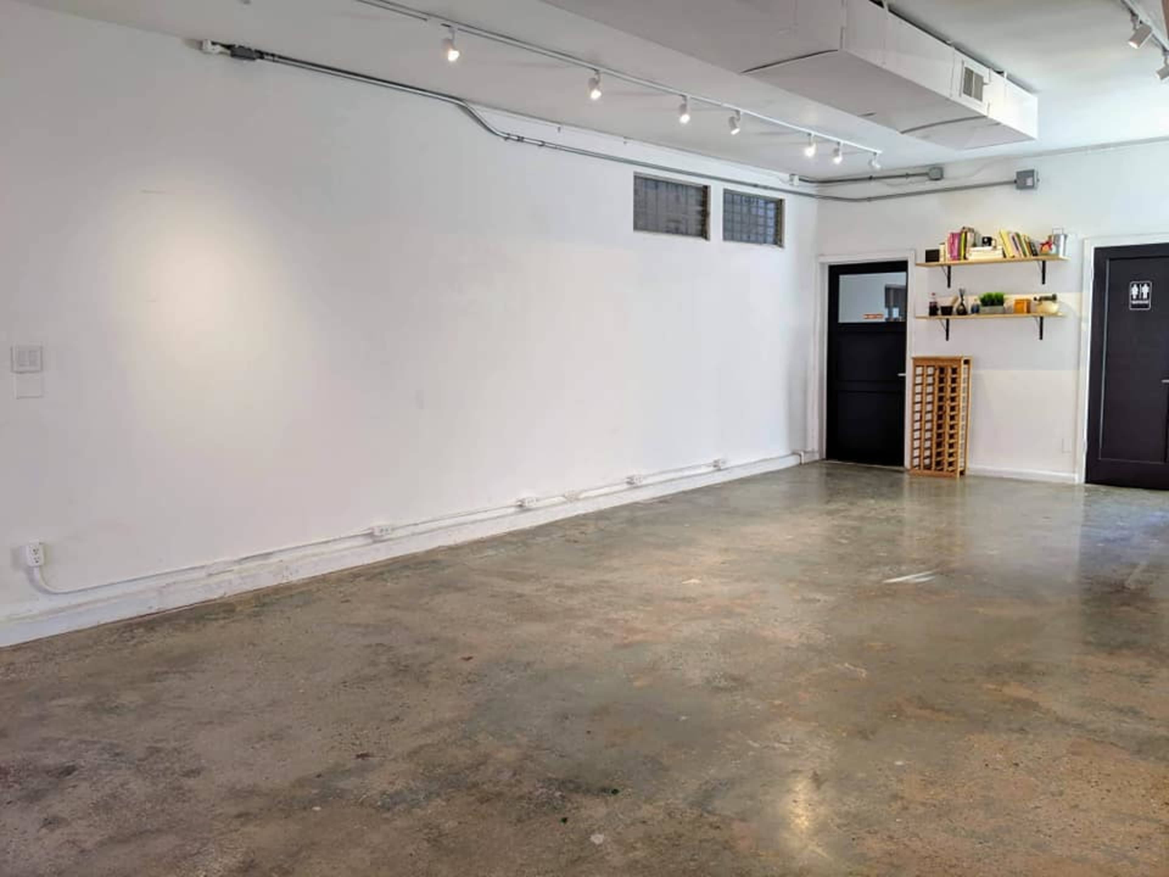 The image shows a spacious, empty room with polished concrete flooring, white walls, and a shelf containing books and decor on one side.