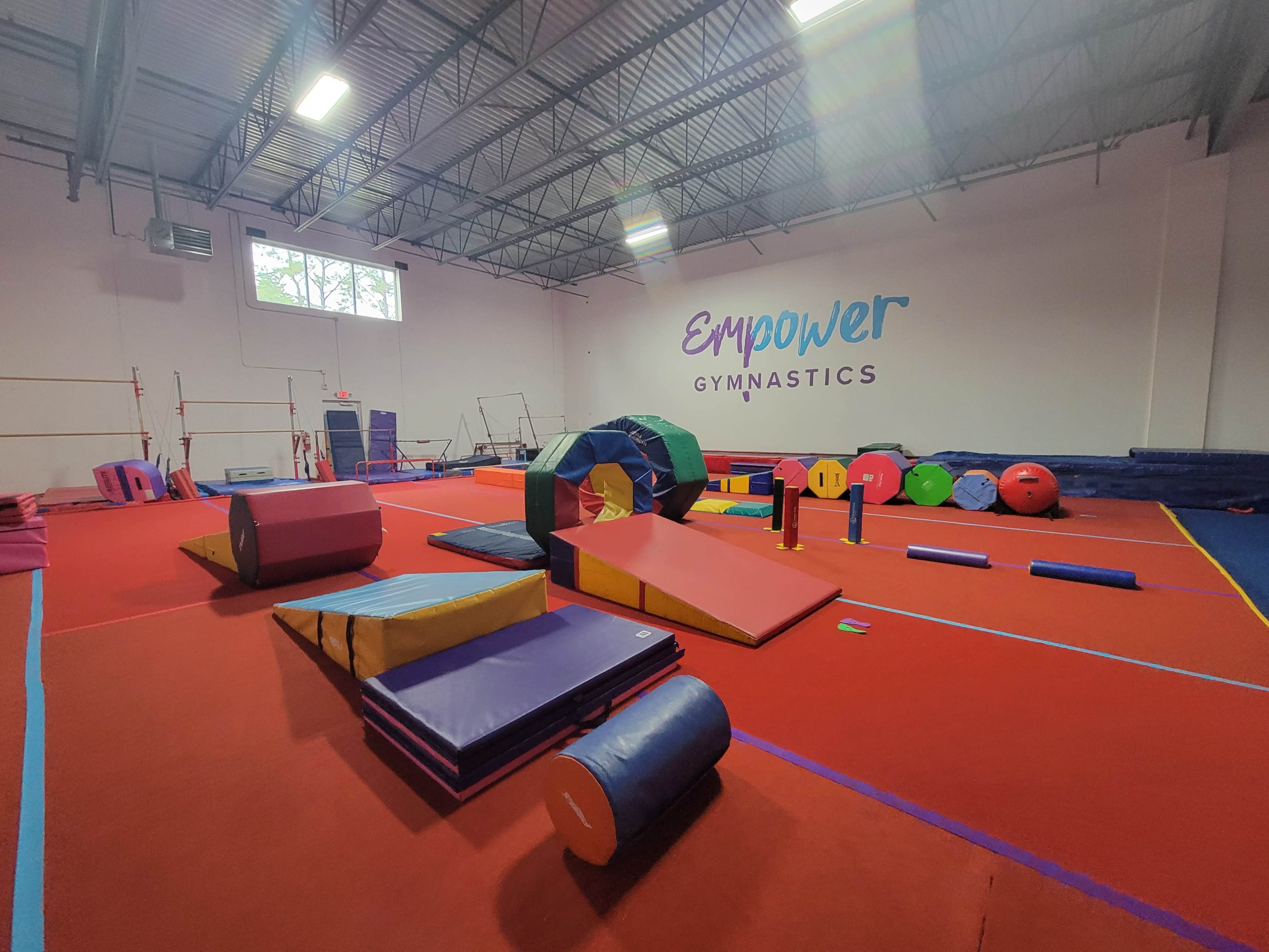 A brightly colored gymnastics facility featuring various equipment and soft mats arranged on a red floor, with the word "Empower" displayed on the wall.