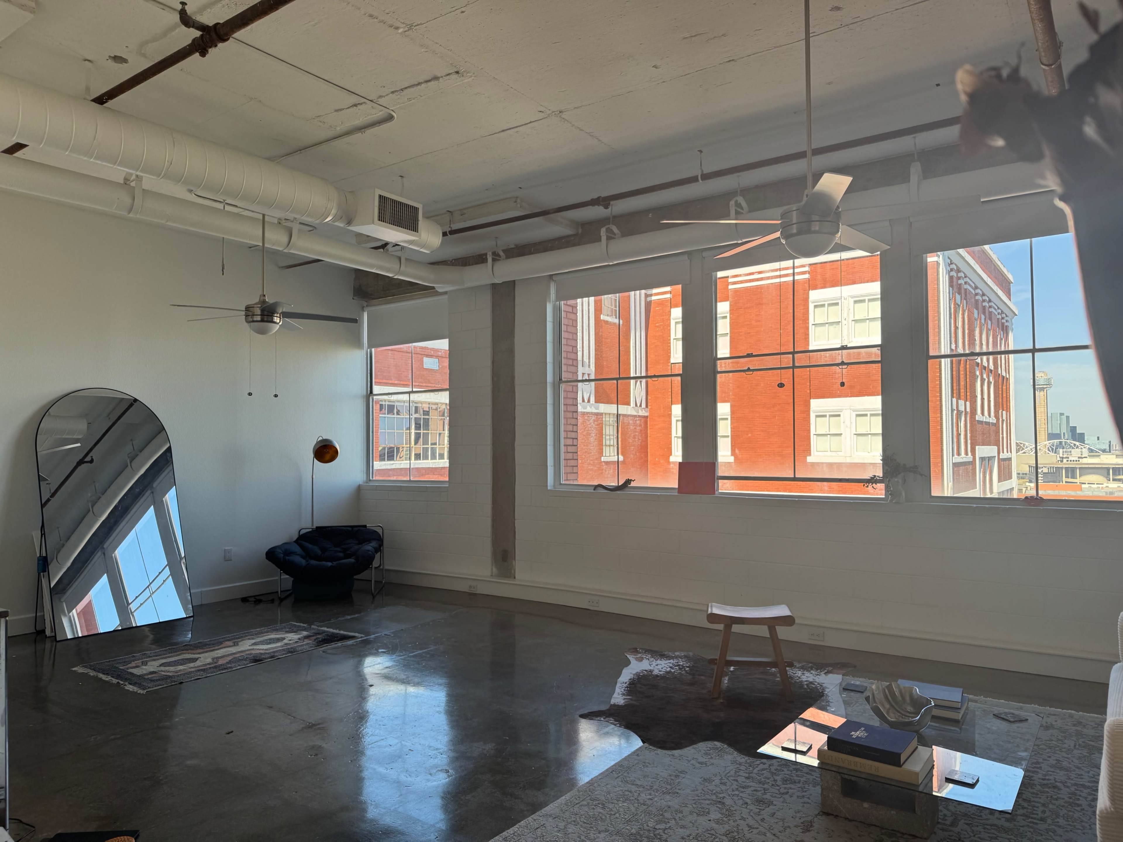 The image shows a spacious room with large windows, concrete floors, and minimal furnishings, including a blue chair, a table, and an abstract rug.