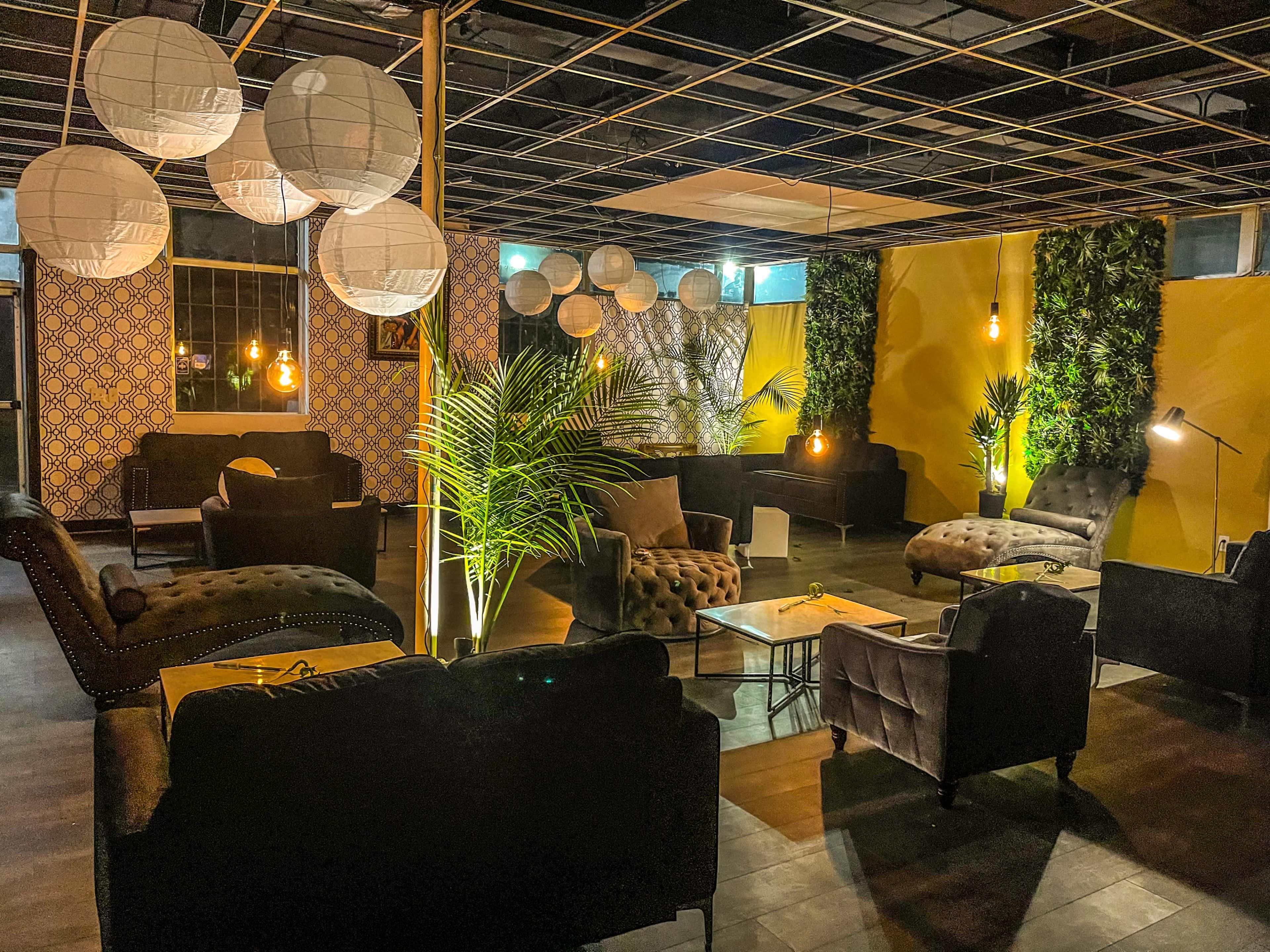 A modern lounge area featuring dark furniture, decorative plants, and soft lighting from pendant lamps and paper lanterns.
