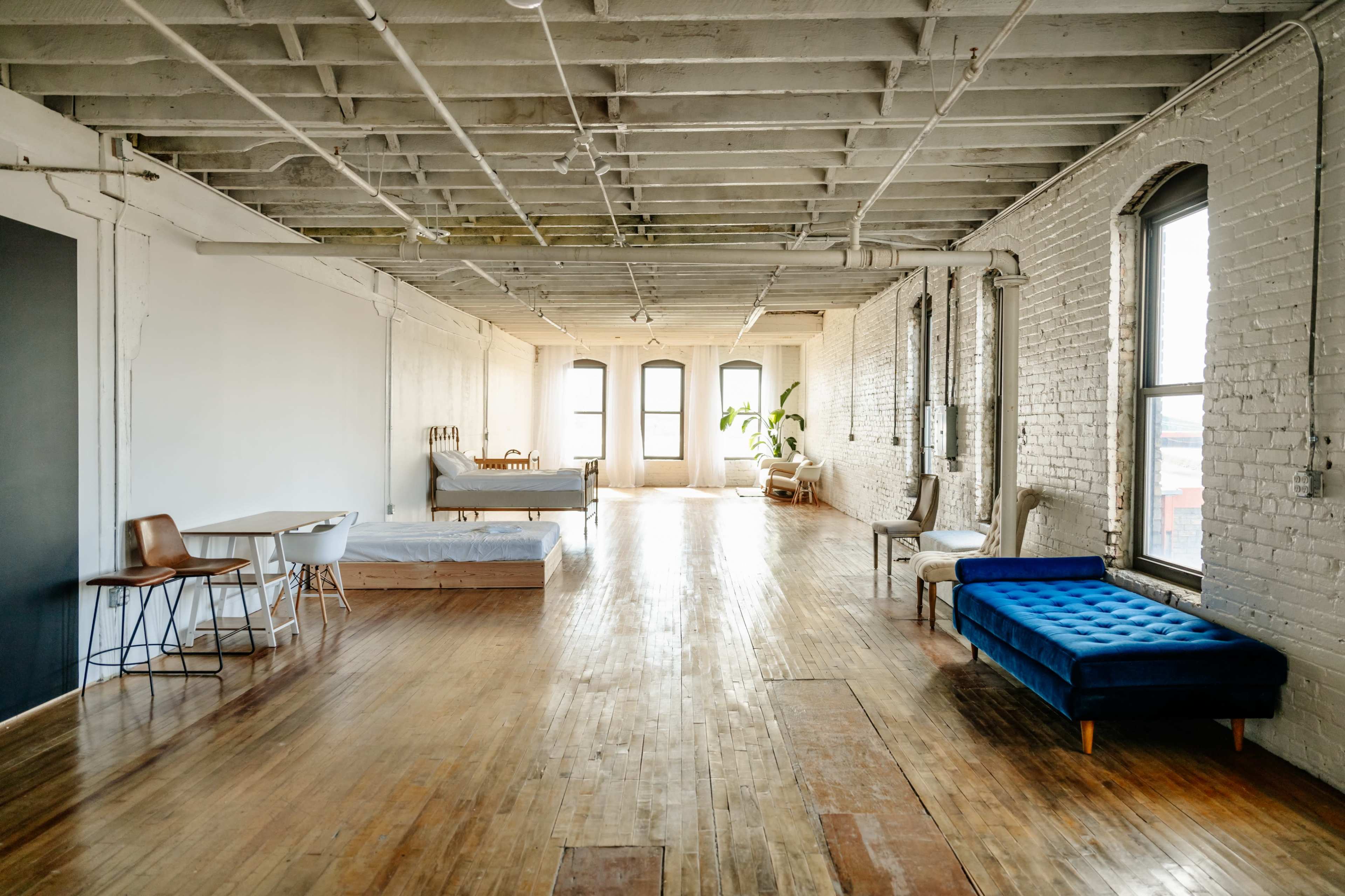 A spacious, minimalist room with wooden floors, large windows, and various furniture pieces, including a bed, a blue couch, and a dining table.