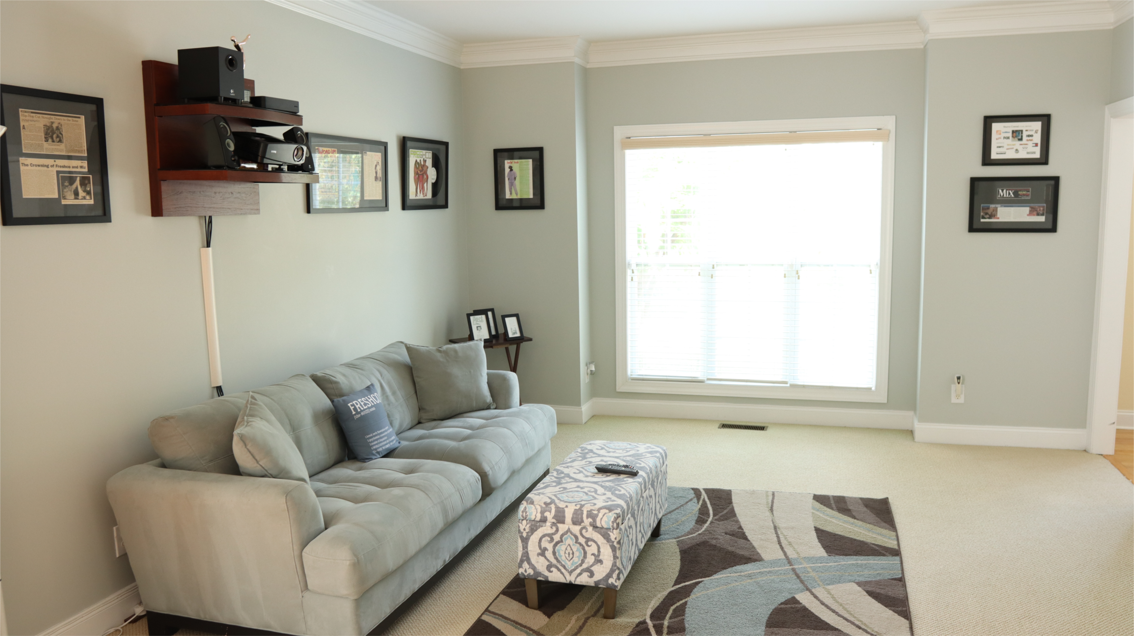 A light gray living room with a sofa, a coffee table on a patterned rug, and framed artwork on the walls, alongside a large window.