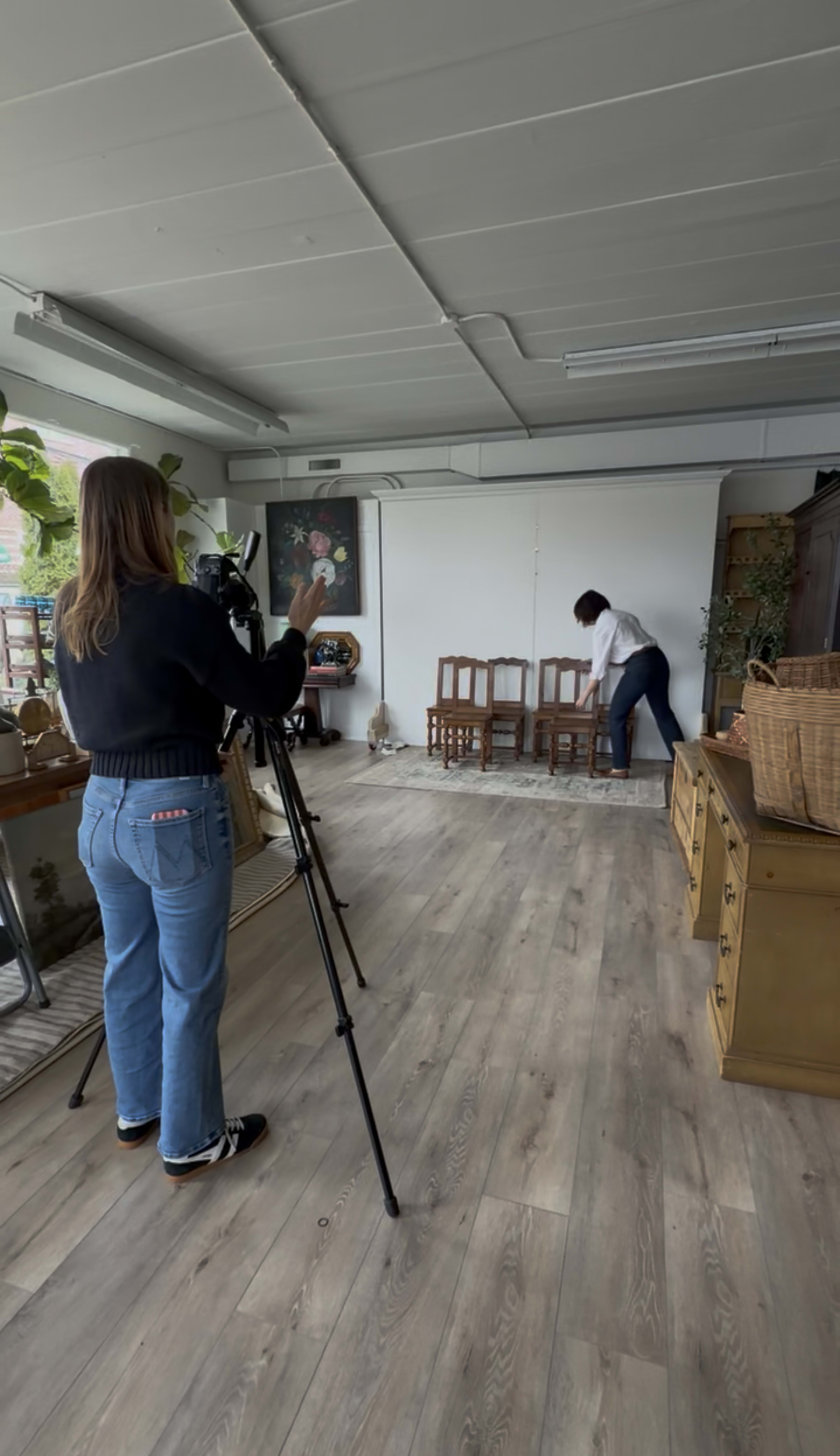 A woman in a white shirt adjusts wooden chairs while another woman with a camera observes in a spacious, well-lit room.