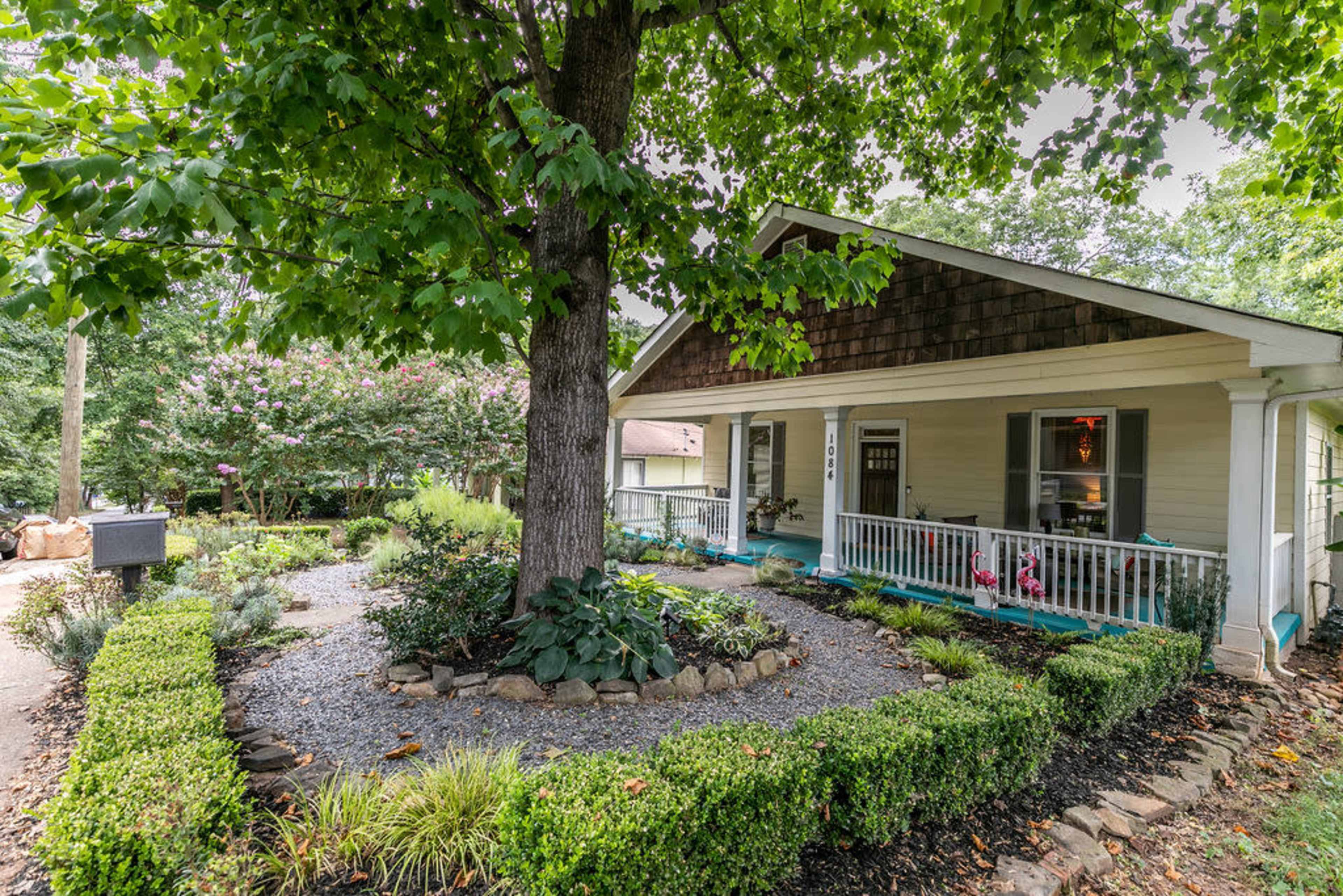 A single-story house with a front porch is surrounded by lush greenery and landscaped flower beds.