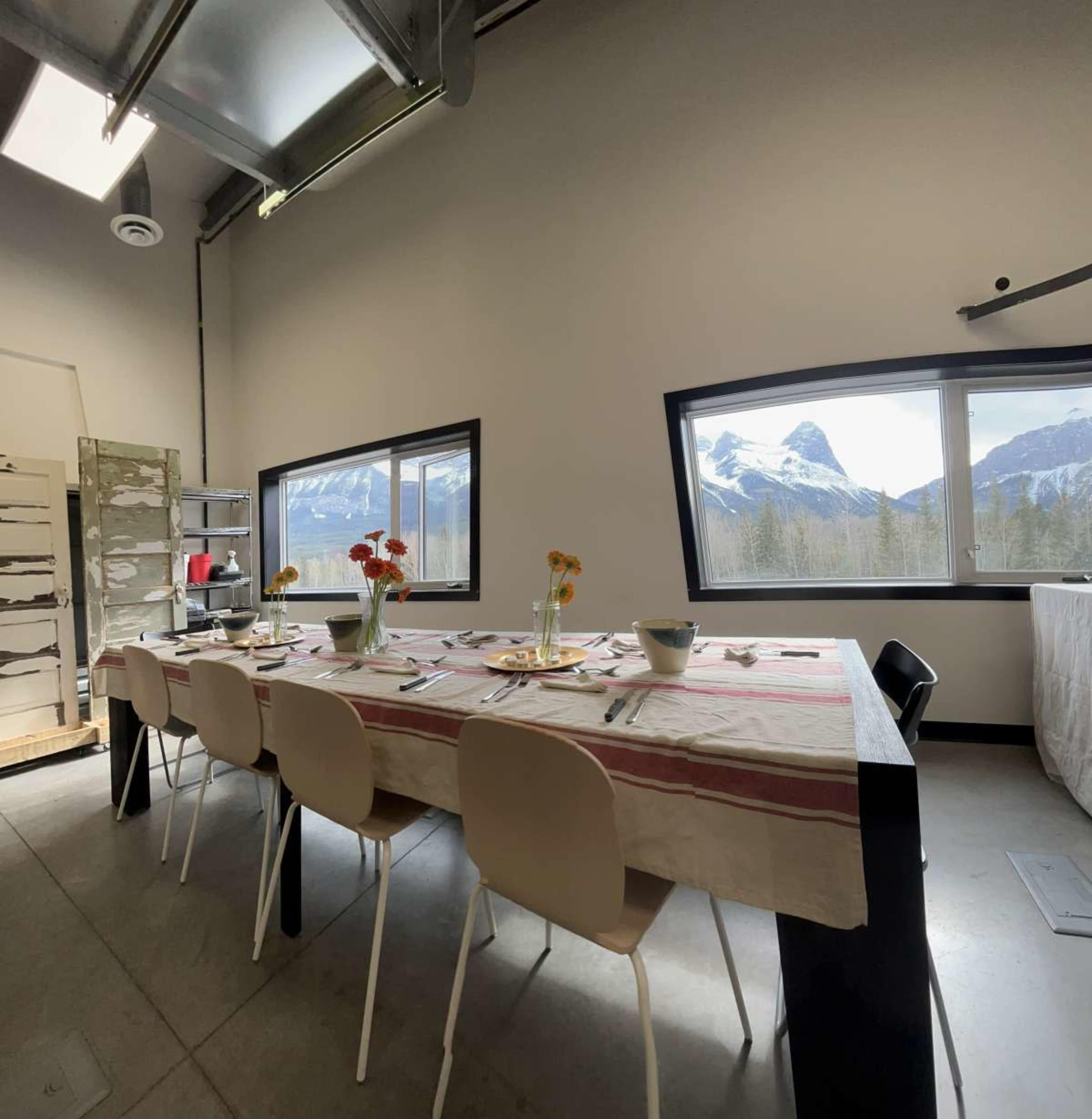 A long dining table is set with utensils and flowers, positioned near large windows that offer a view of mountains.