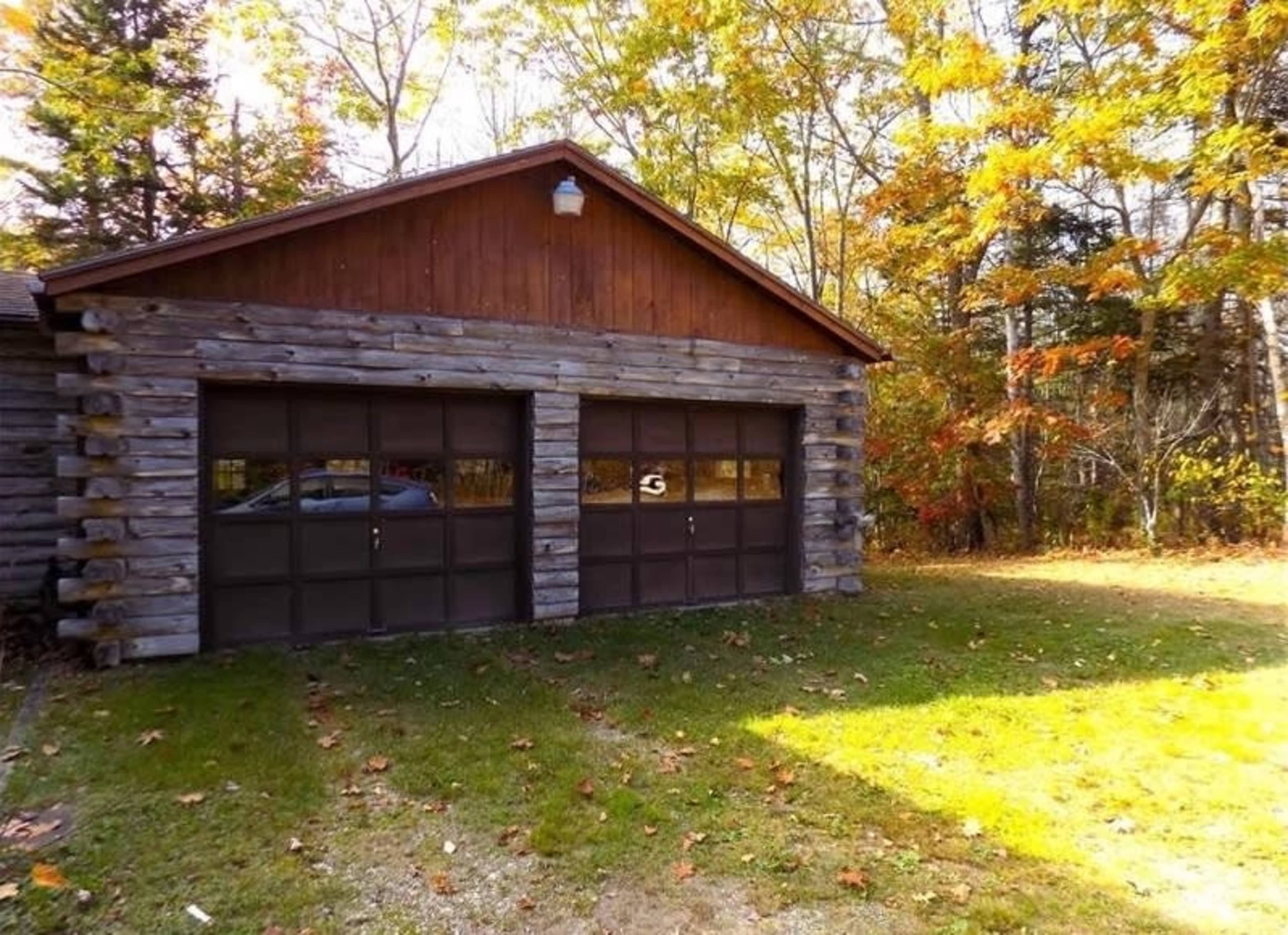 A log-style garage with two doors, set in a grassy area surrounded by autumn-colored trees.