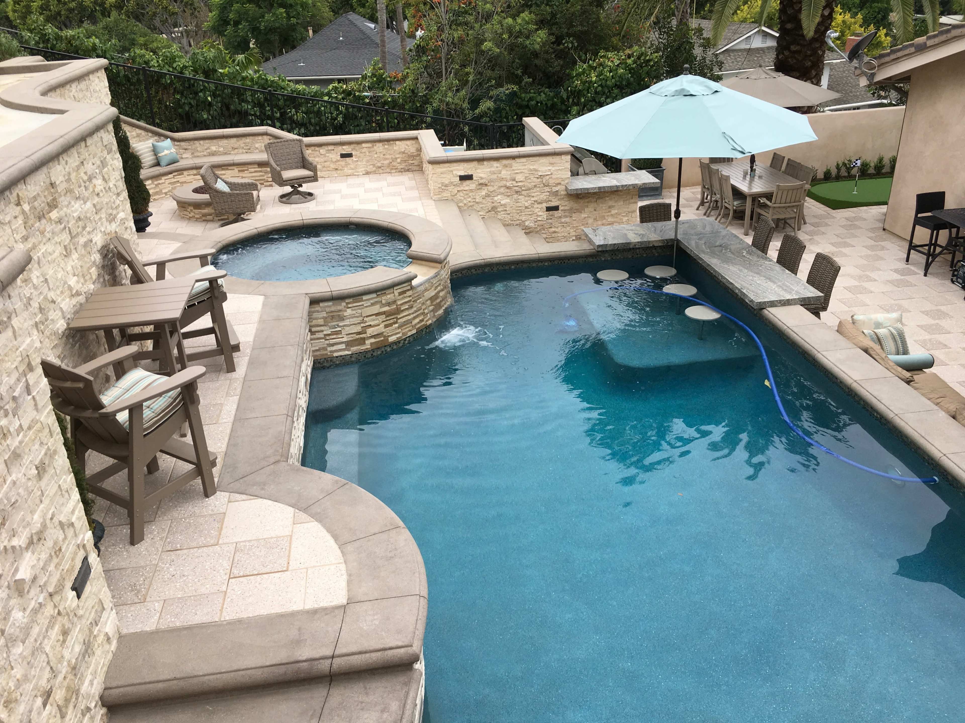 The image shows a spacious backyard with a swimming pool, a hot tub, outdoor seating, and a large umbrella for shade.