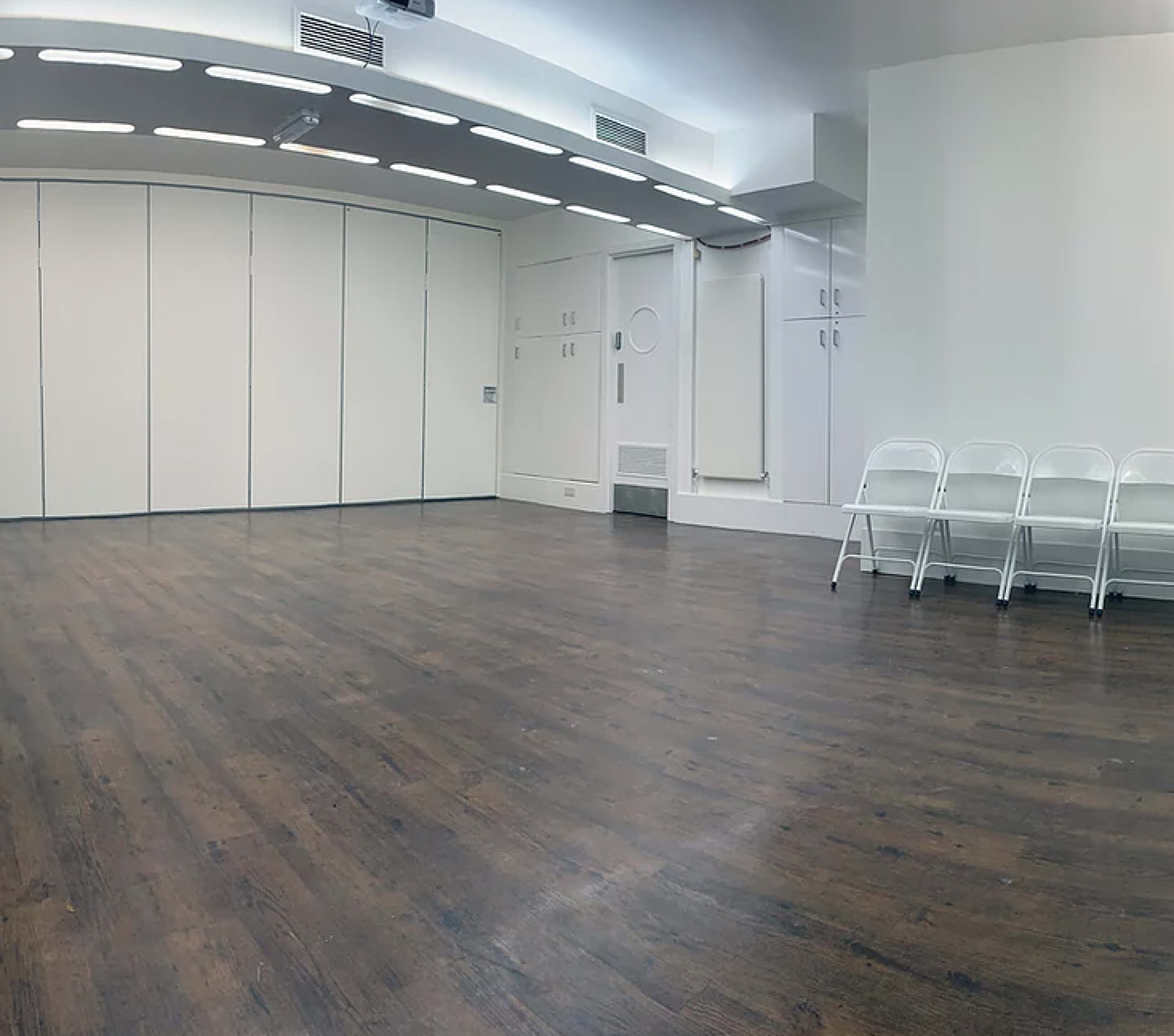 A spacious, empty room with a polished wooden floor and several white folding chairs arranged against a wall.