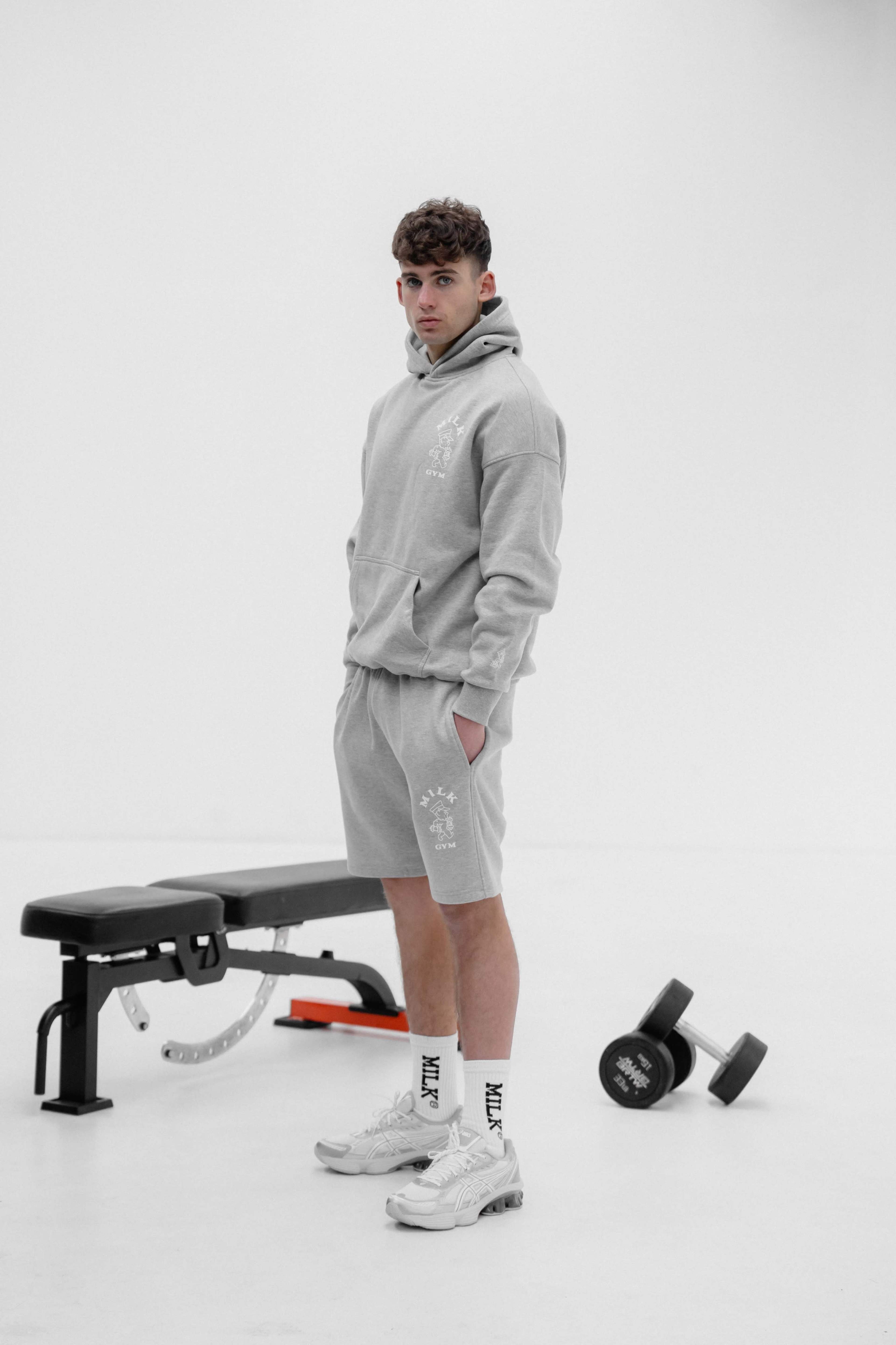 A young man in a gray hoodie and shorts stands next to a weight bench and dumbbell in a minimalistic gym setting.