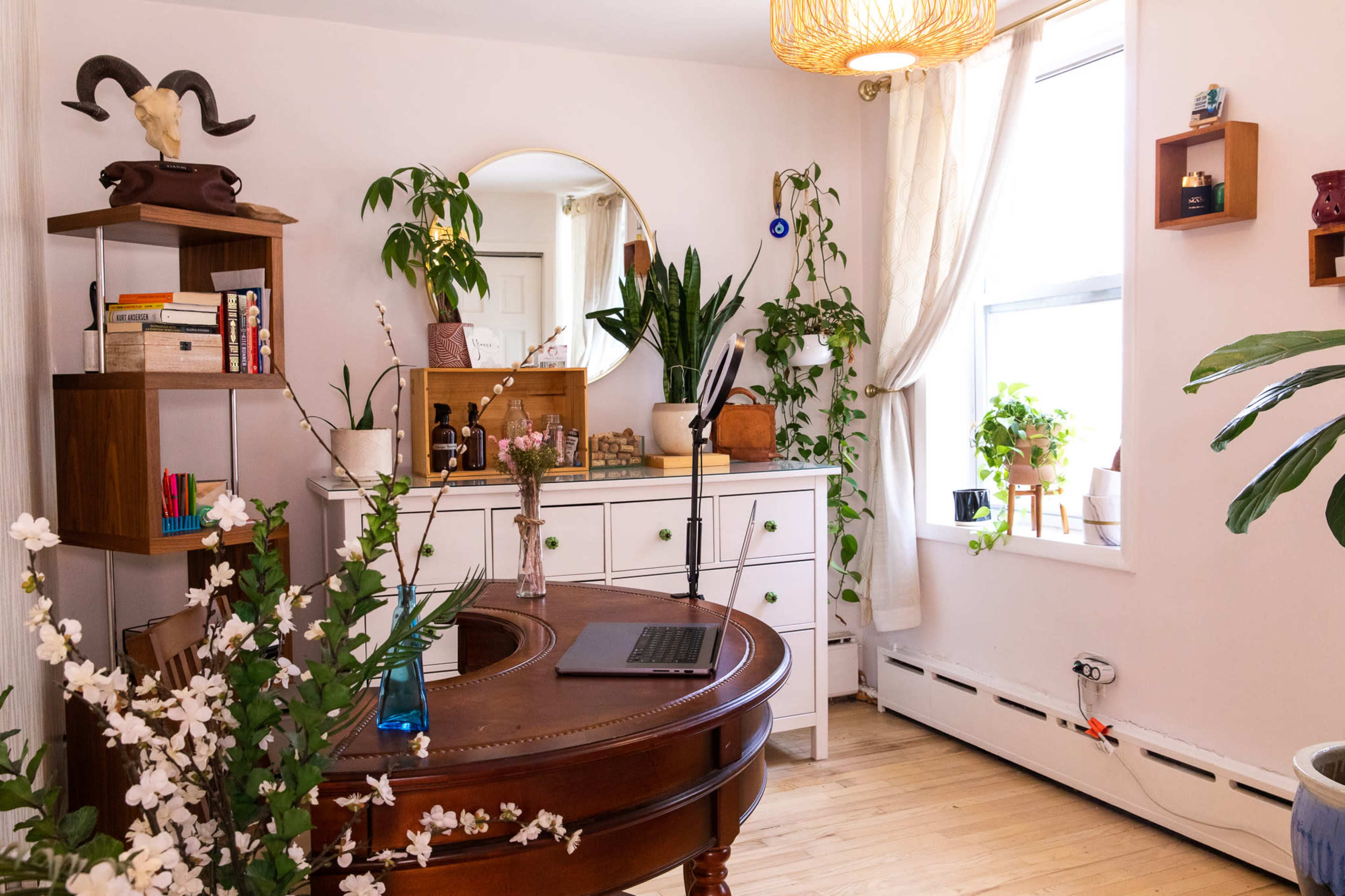 The image shows a well-decorated home office featuring a wooden desk, a dresser, several plants, and a round mirror on the wall.