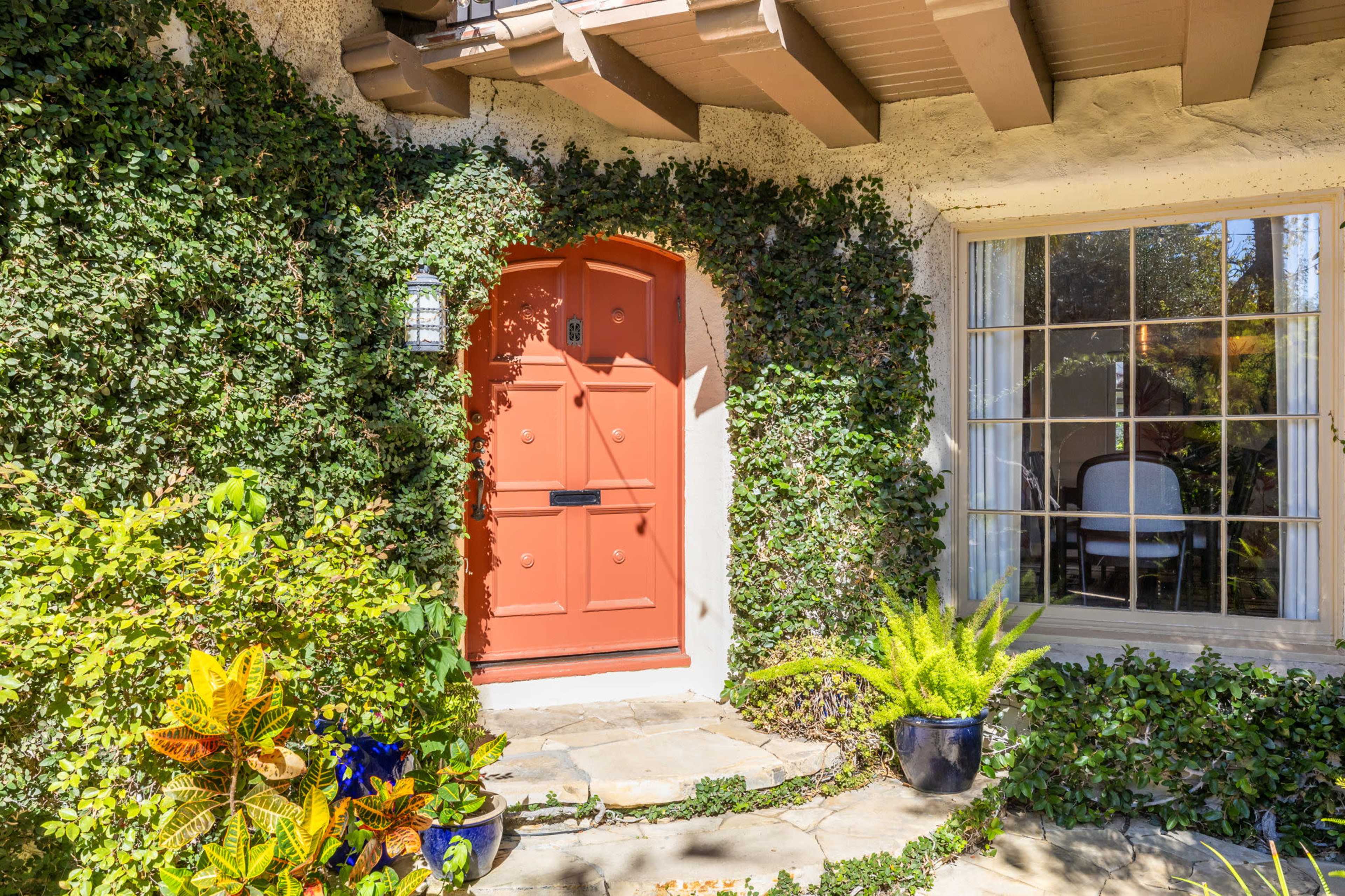 A decorative orange door is framed by green ivy and surrounded by various plants next to a window with white curtains.