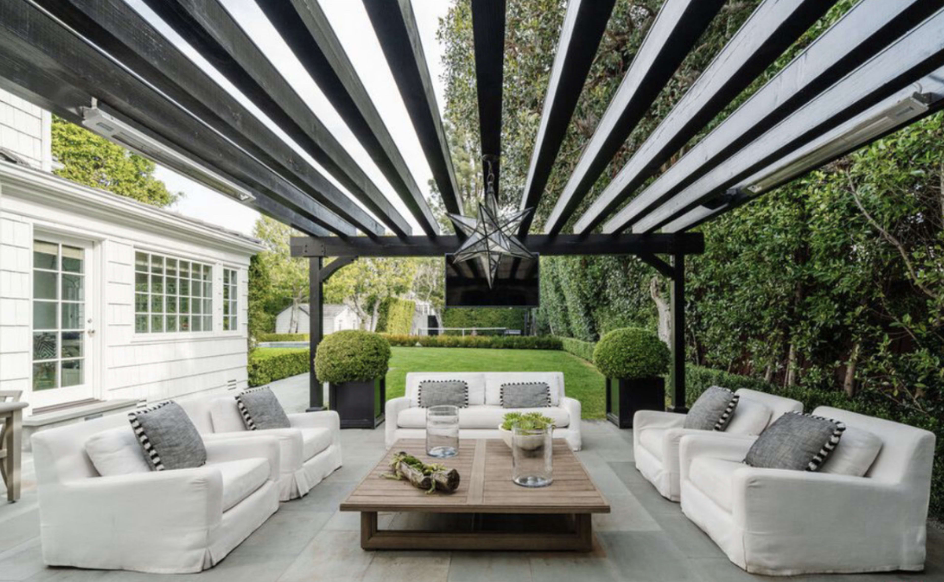 A modern outdoor patio features white sofas arranged around a wooden coffee table, shaded by a slatted pergola with green hedges in the background.