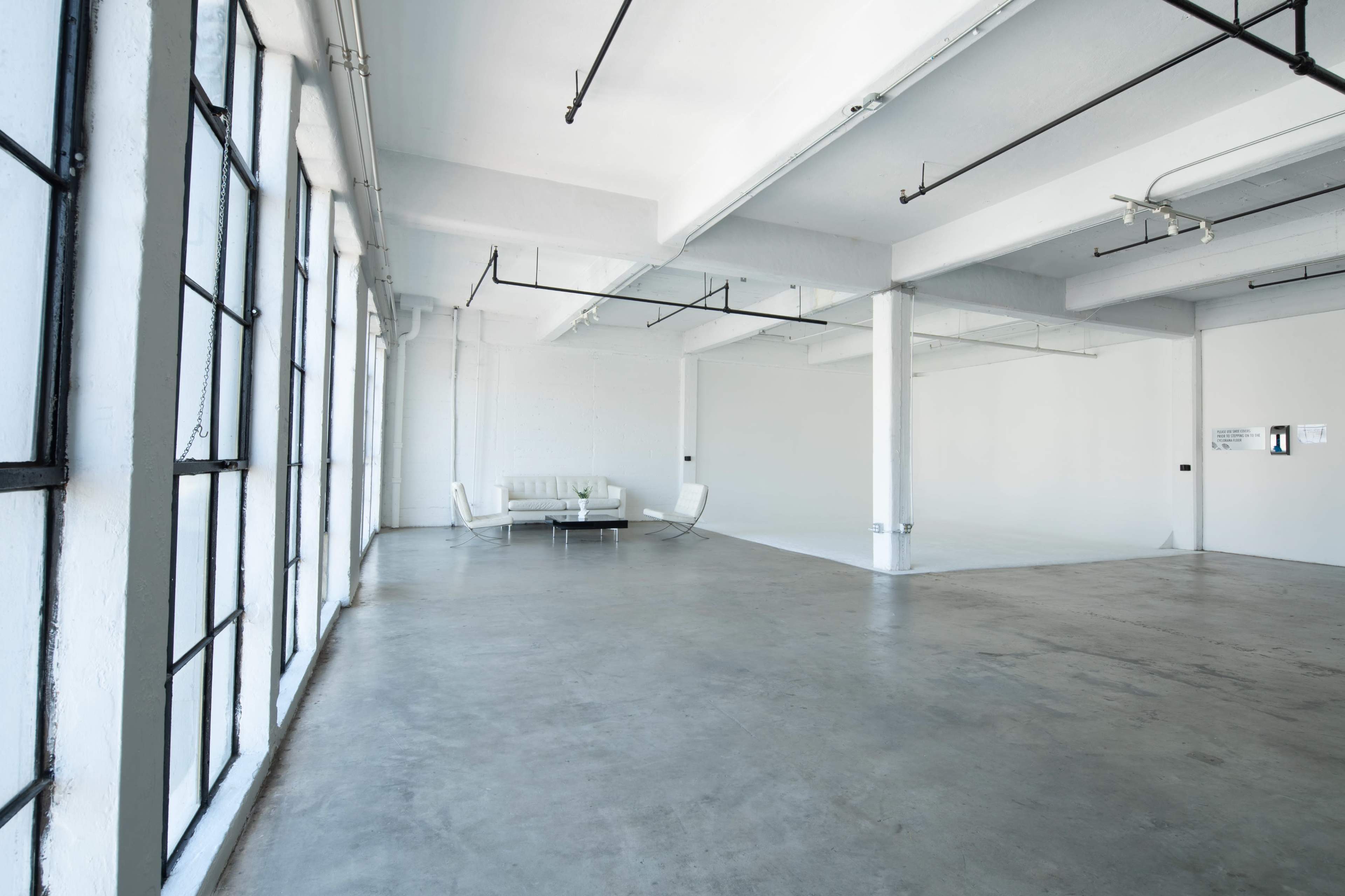 A spacious, minimalistic room with concrete flooring and large windows, featuring a white sofa and chairs.