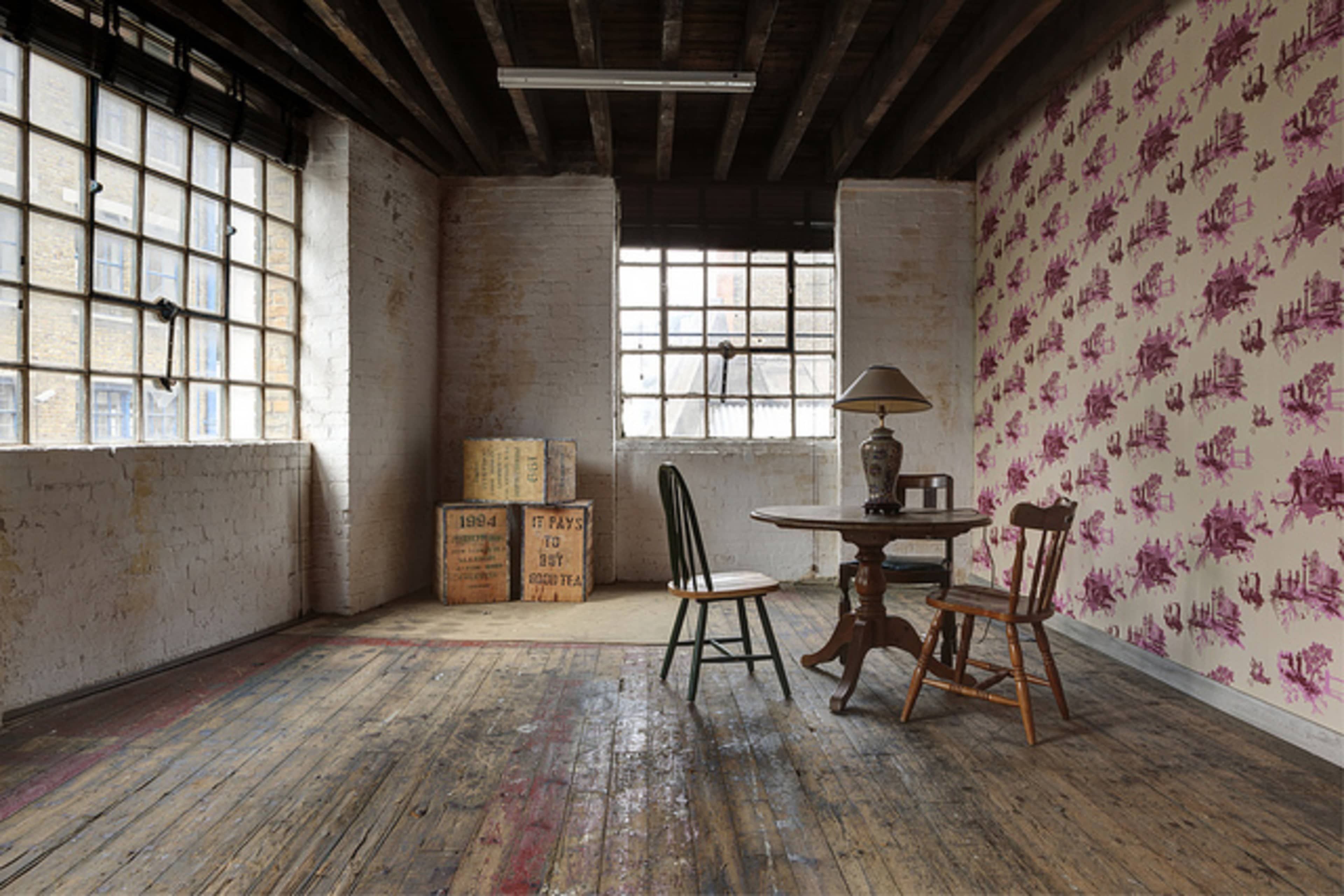 An empty room features a wooden table with two chairs, a lamp, and boxes stacked against a wall adorned with floral wallpaper.
