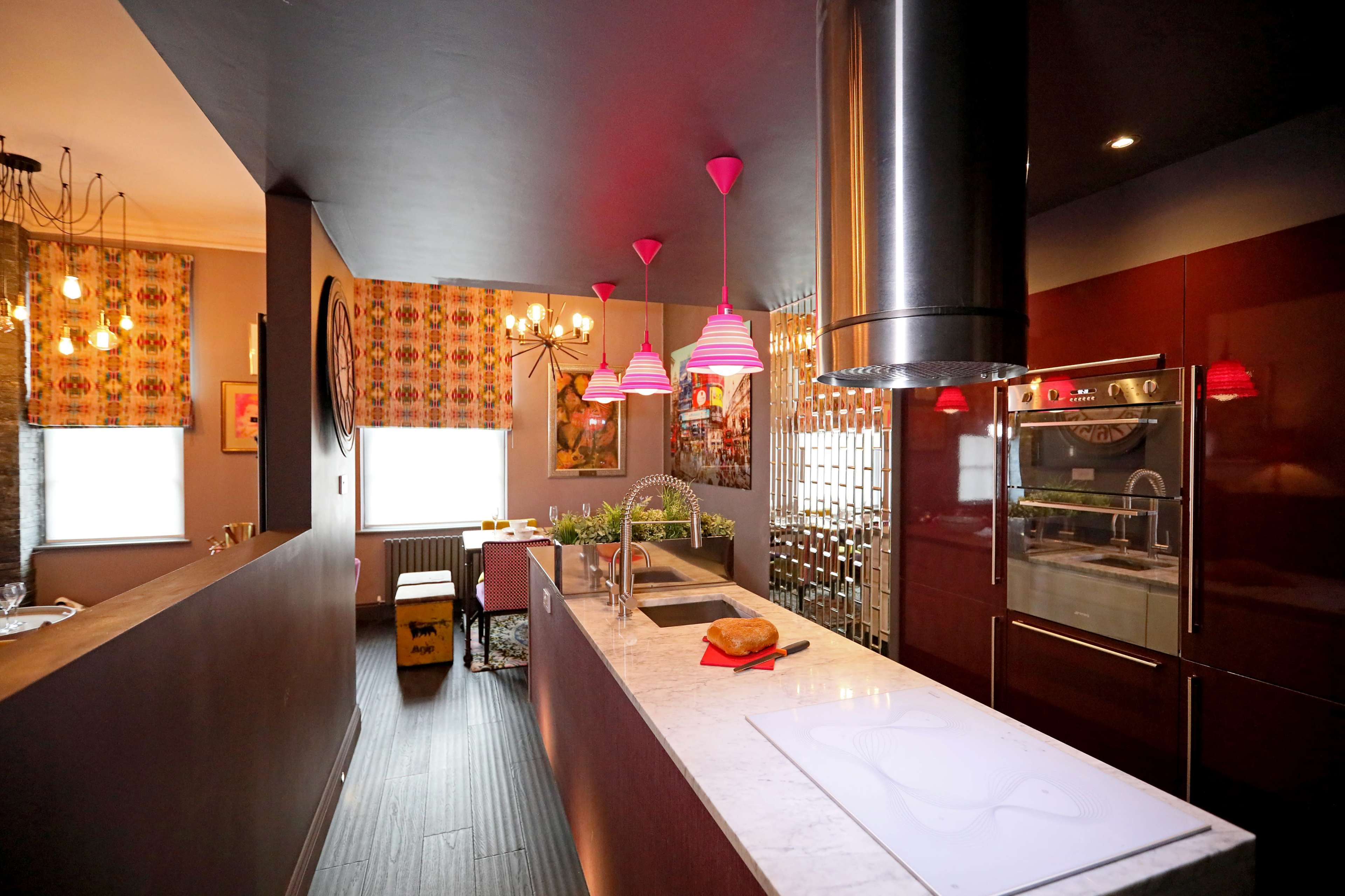 A modern kitchen with dark cabinetry, a marble countertop, pink hanging light fixtures, and an island with a stovetop, complemented by decorative window treatments.
