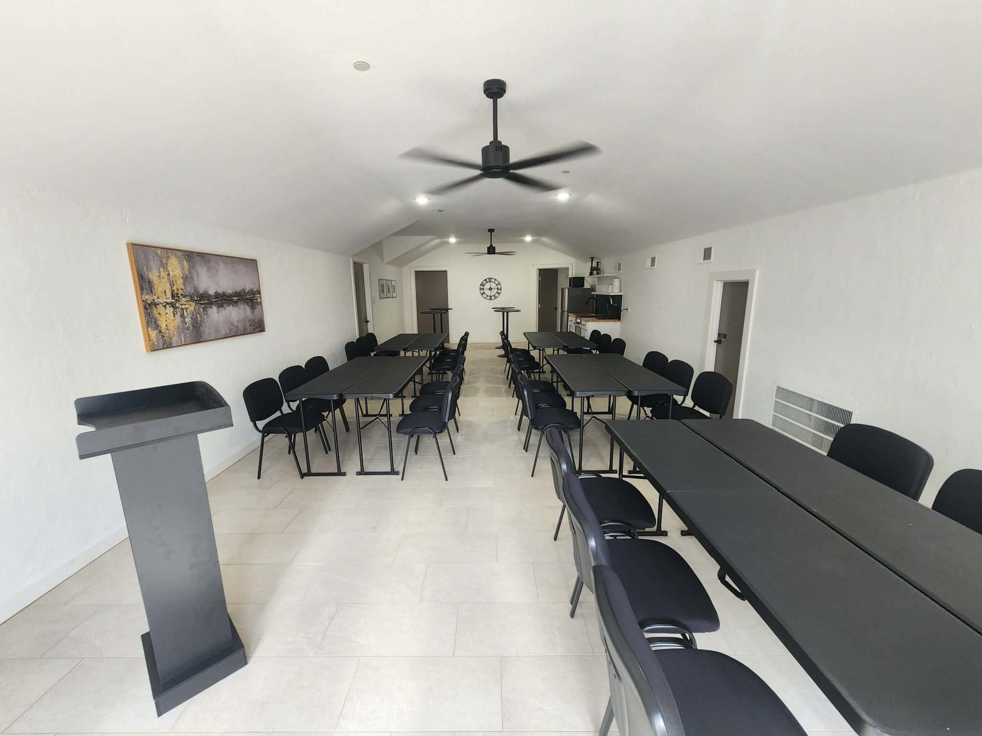 The image shows a spacious meeting room with multiple black tables arranged in rows, surrounded by black chairs, and featuring a podium at the front.
