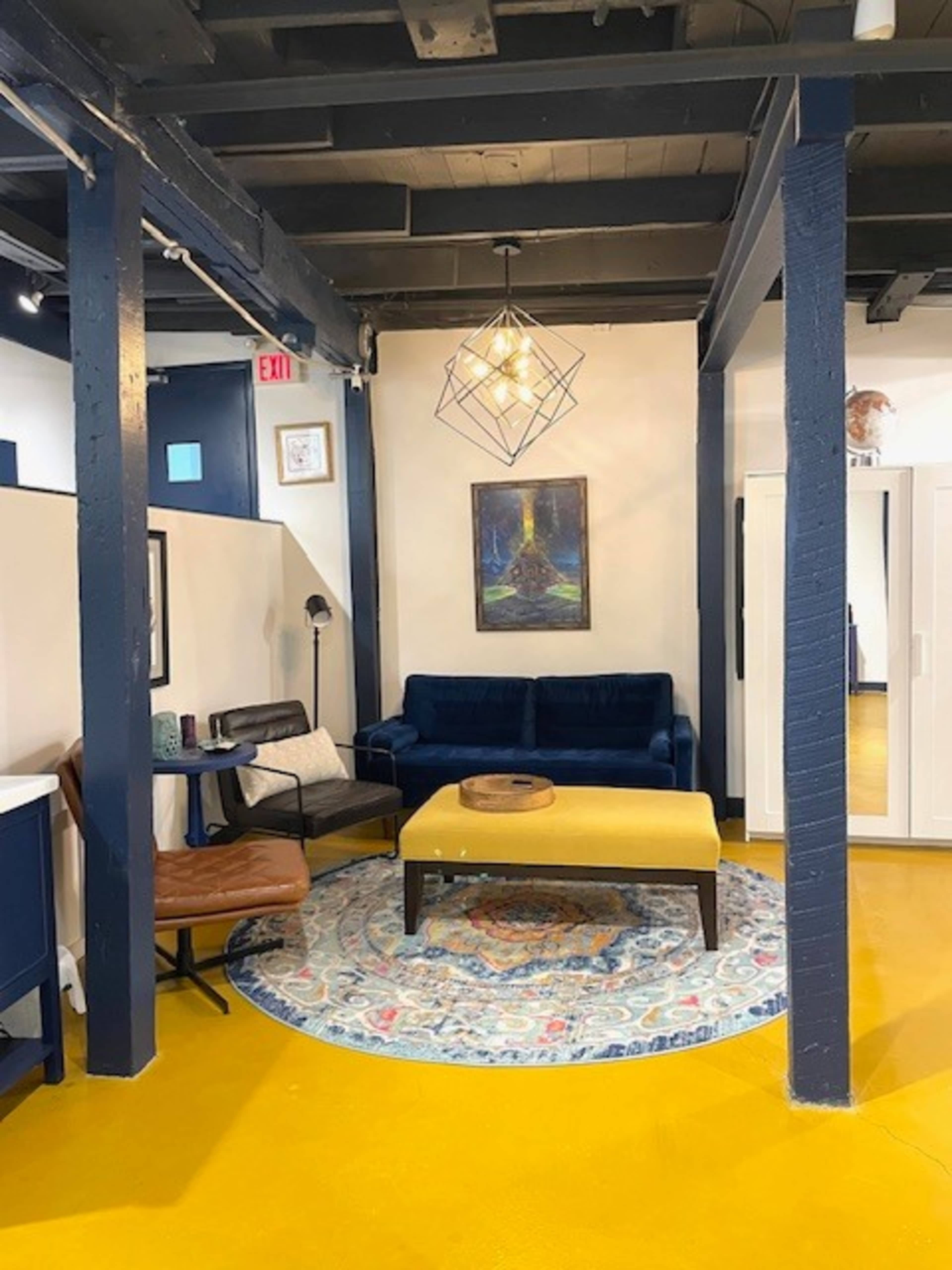 The image shows a modern living area featuring a blue sofa, a yellow coffee table, and an area rug on a yellow floor, with a light fixture and artwork above.