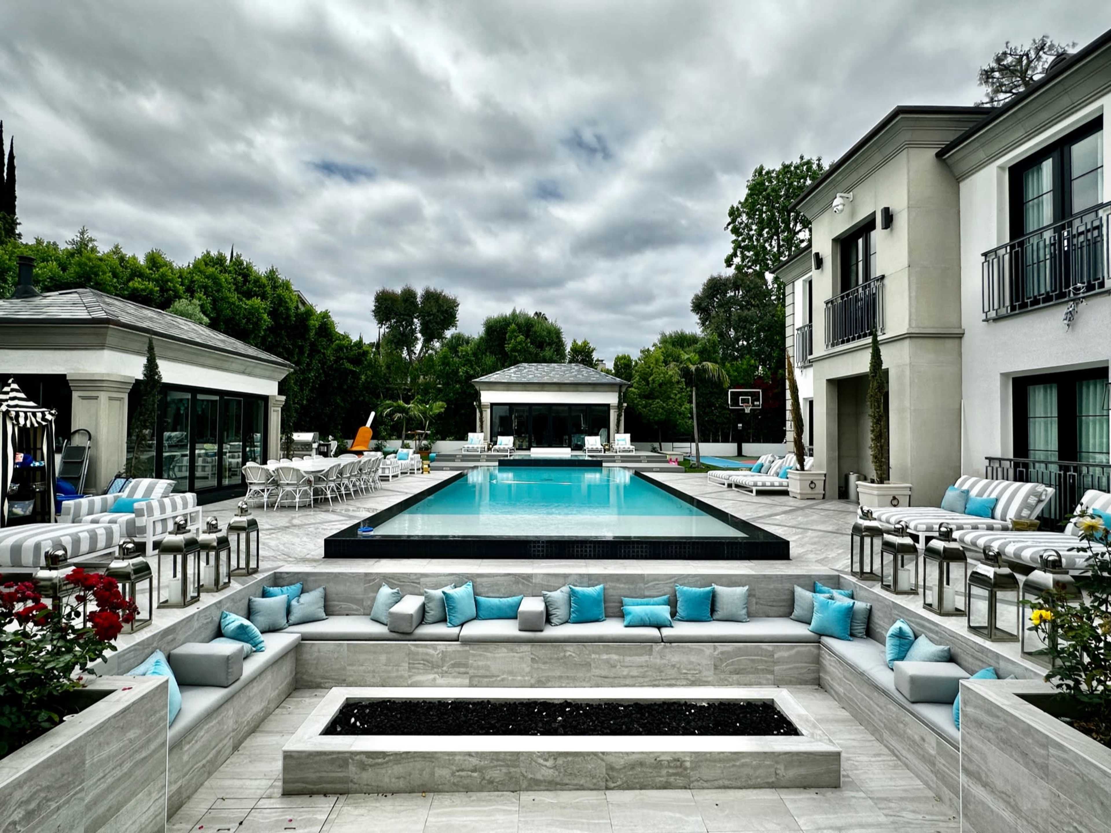 A modern outdoor swimming pool surrounded by lounge chairs, decorative cushions, and a pavilion in a landscaped setting under a cloudy sky.