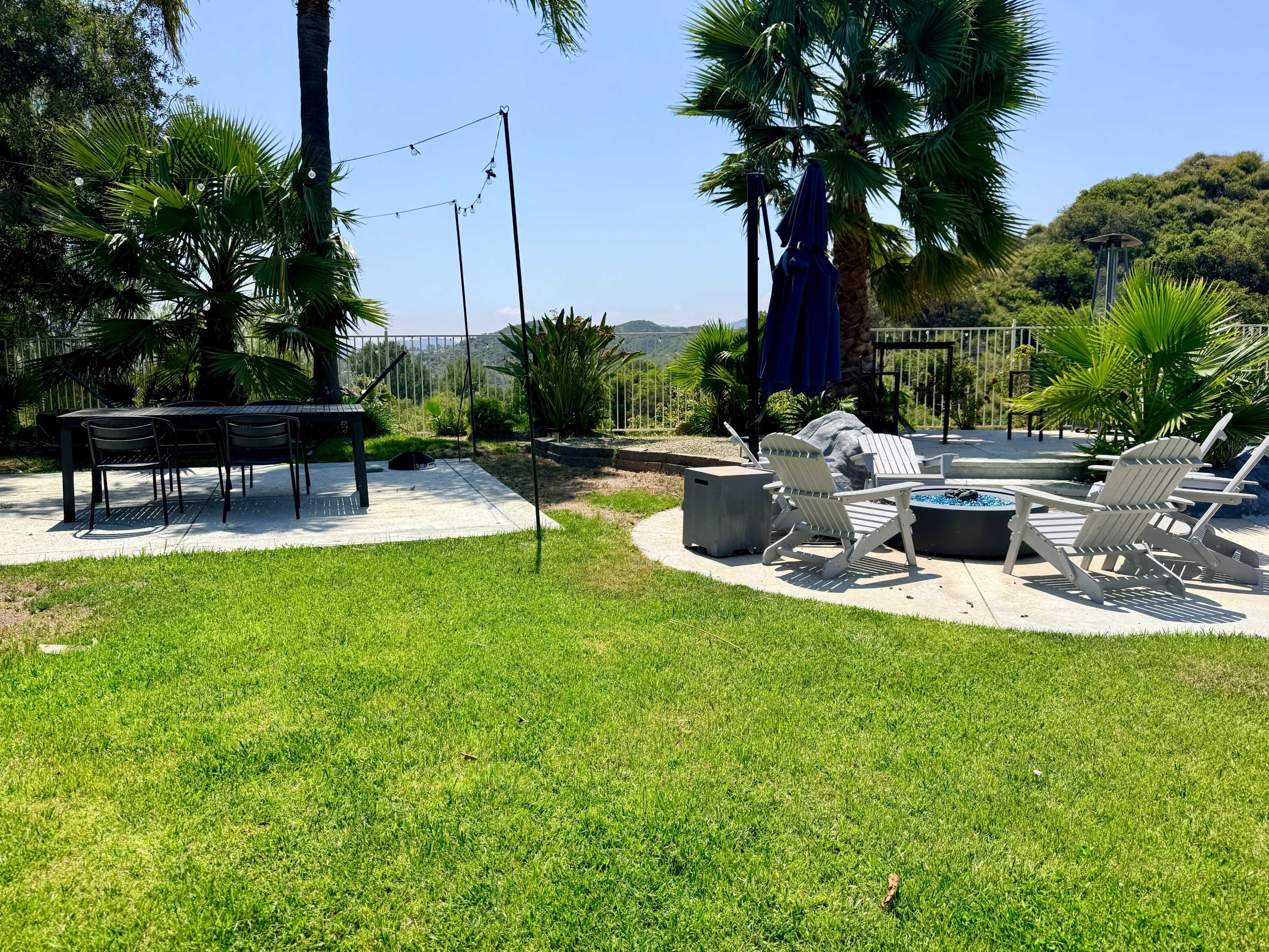 The image shows a landscaped outdoor area with a grassy lawn, dining table, fire pit, and palm trees, set against a hilly background.