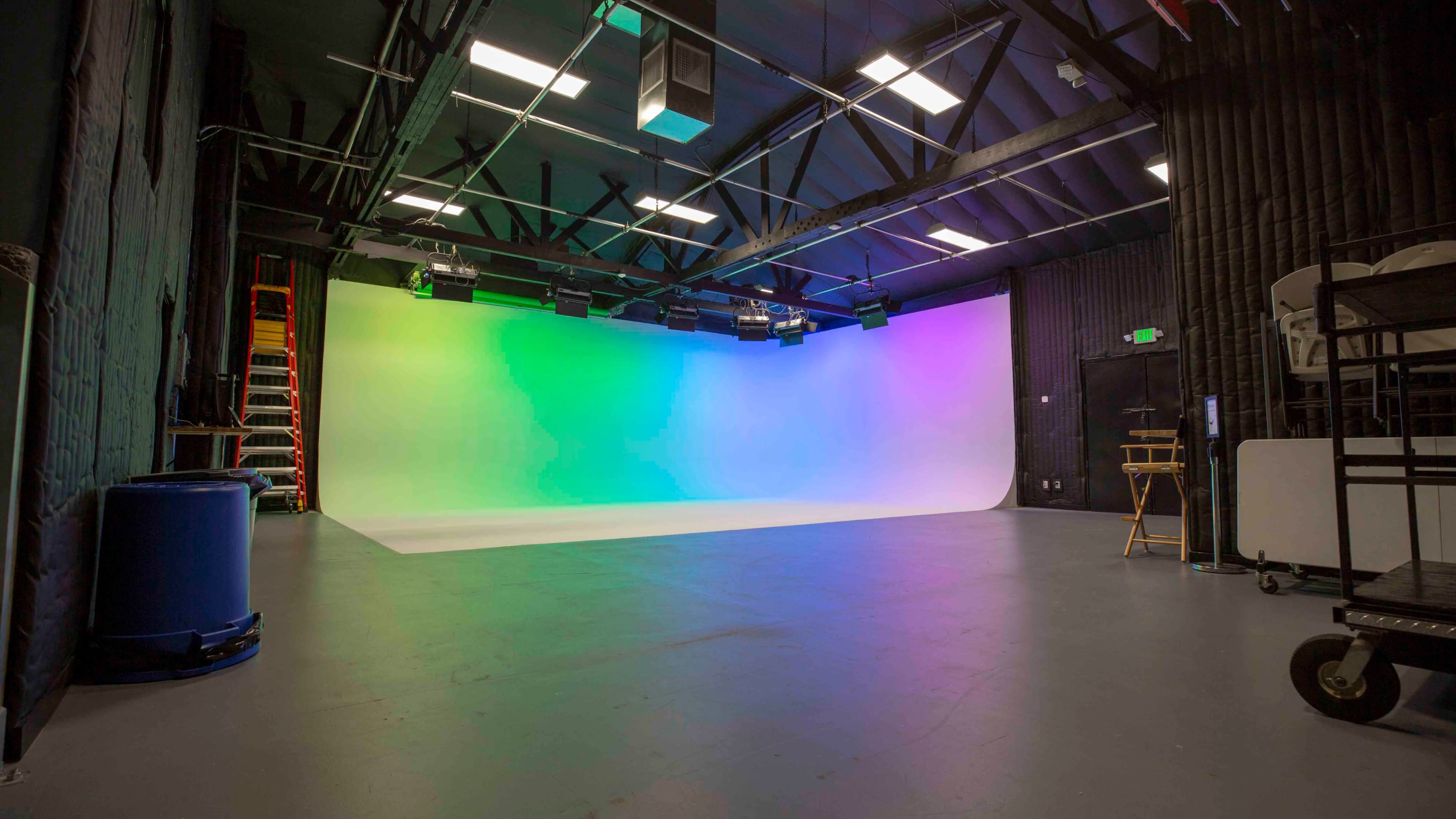 A photo studio features a gradient backdrop of green and purple light, surrounded by black walls and various equipment.