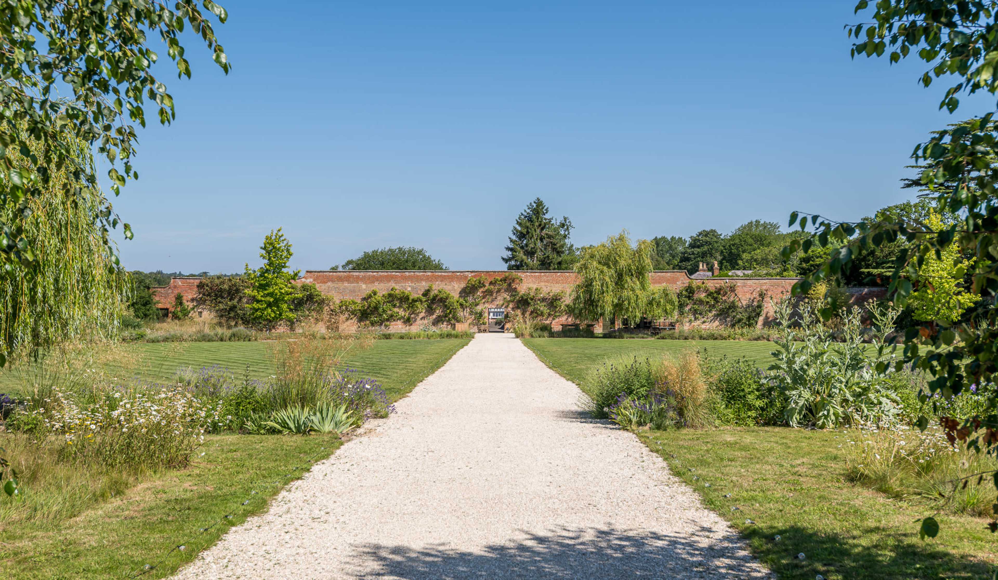 A gravel pathway leads through a landscaped garden toward a brick wall, framed by trees and greenery under a clear blue sky.