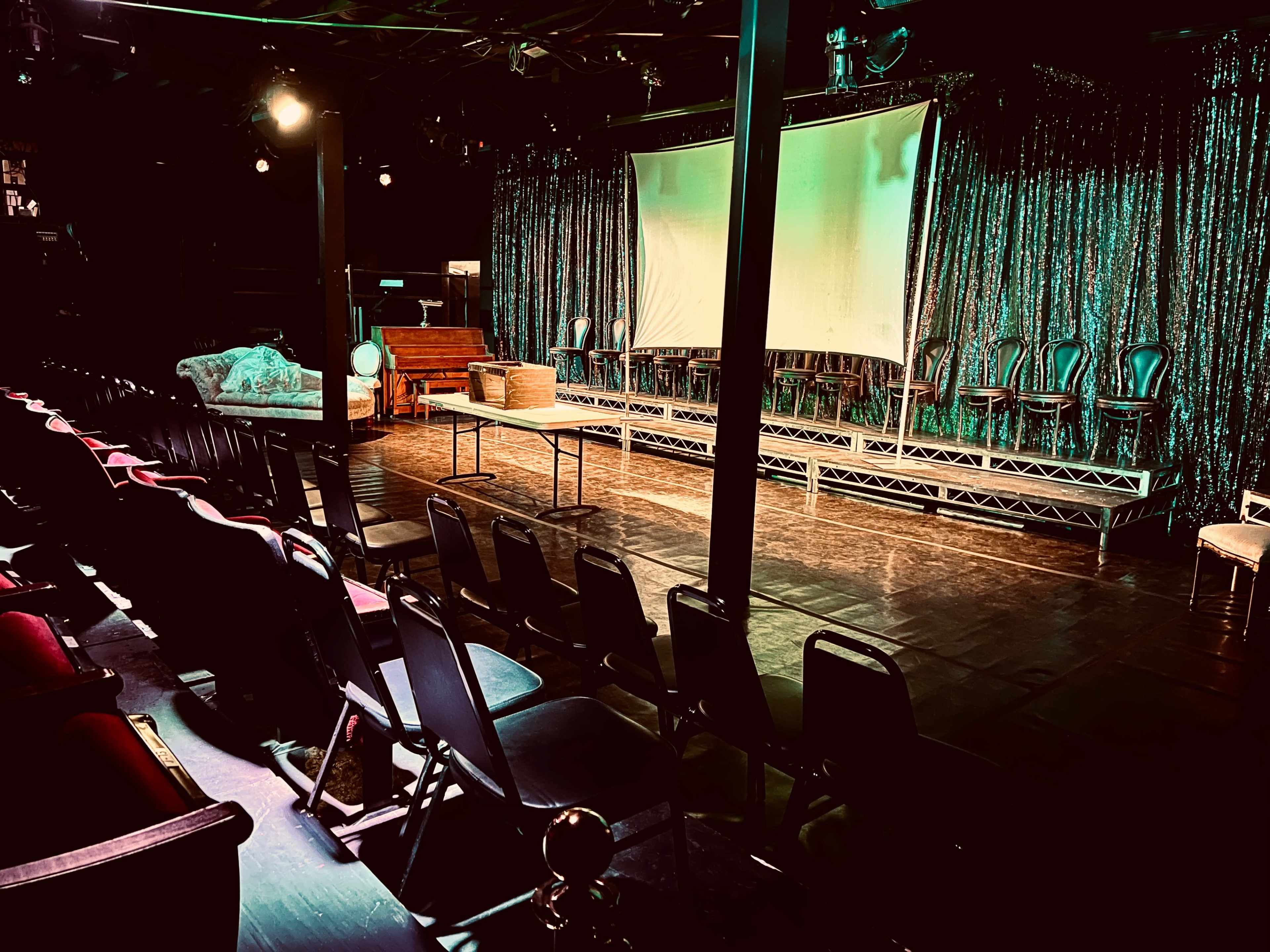 A performance space with rows of chairs facing a stage that has a large screen and a glittering curtain backdrop.