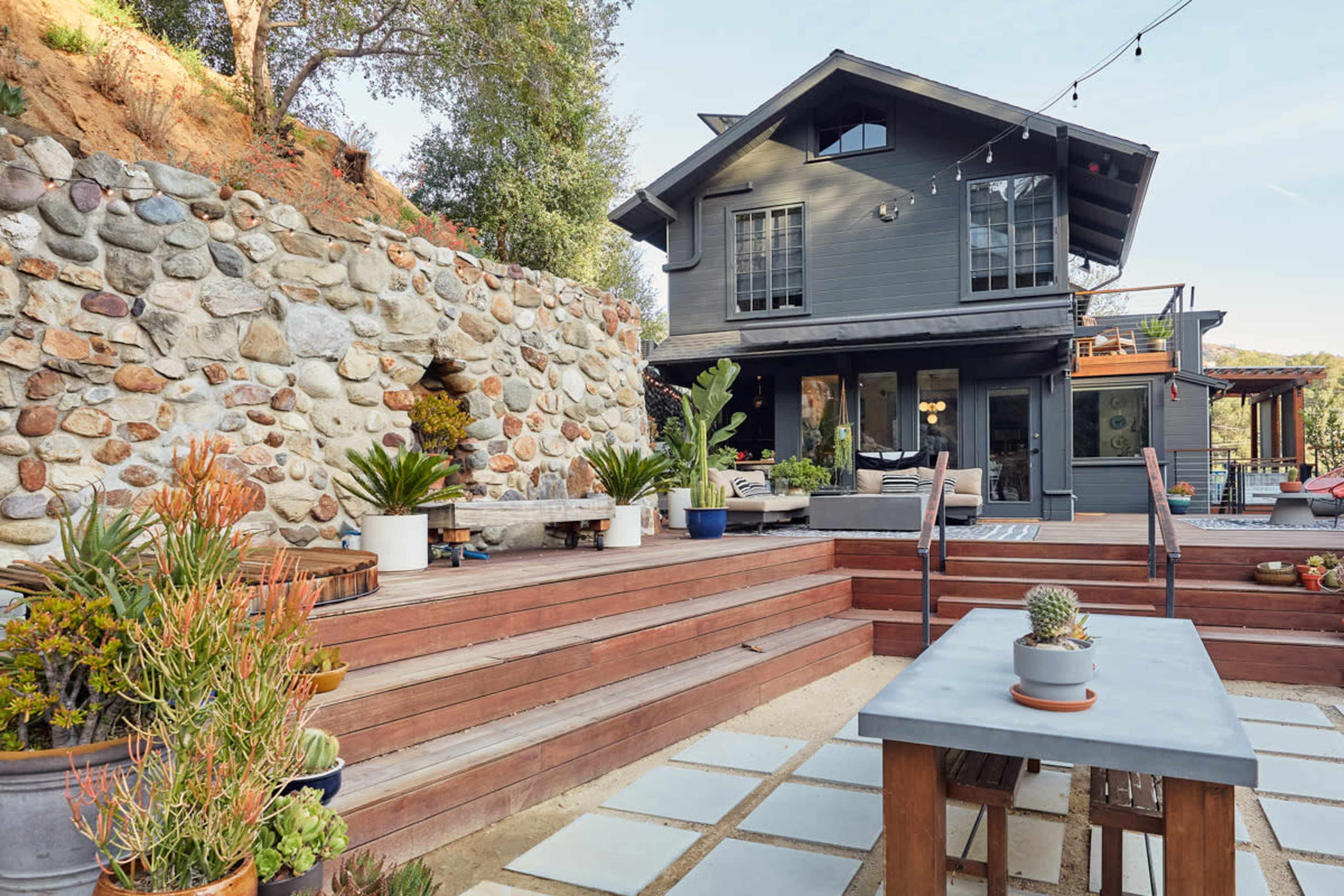 A modern house with a stone wall and wooden steps leading to a patio adorned with potted plants and outdoor furniture.