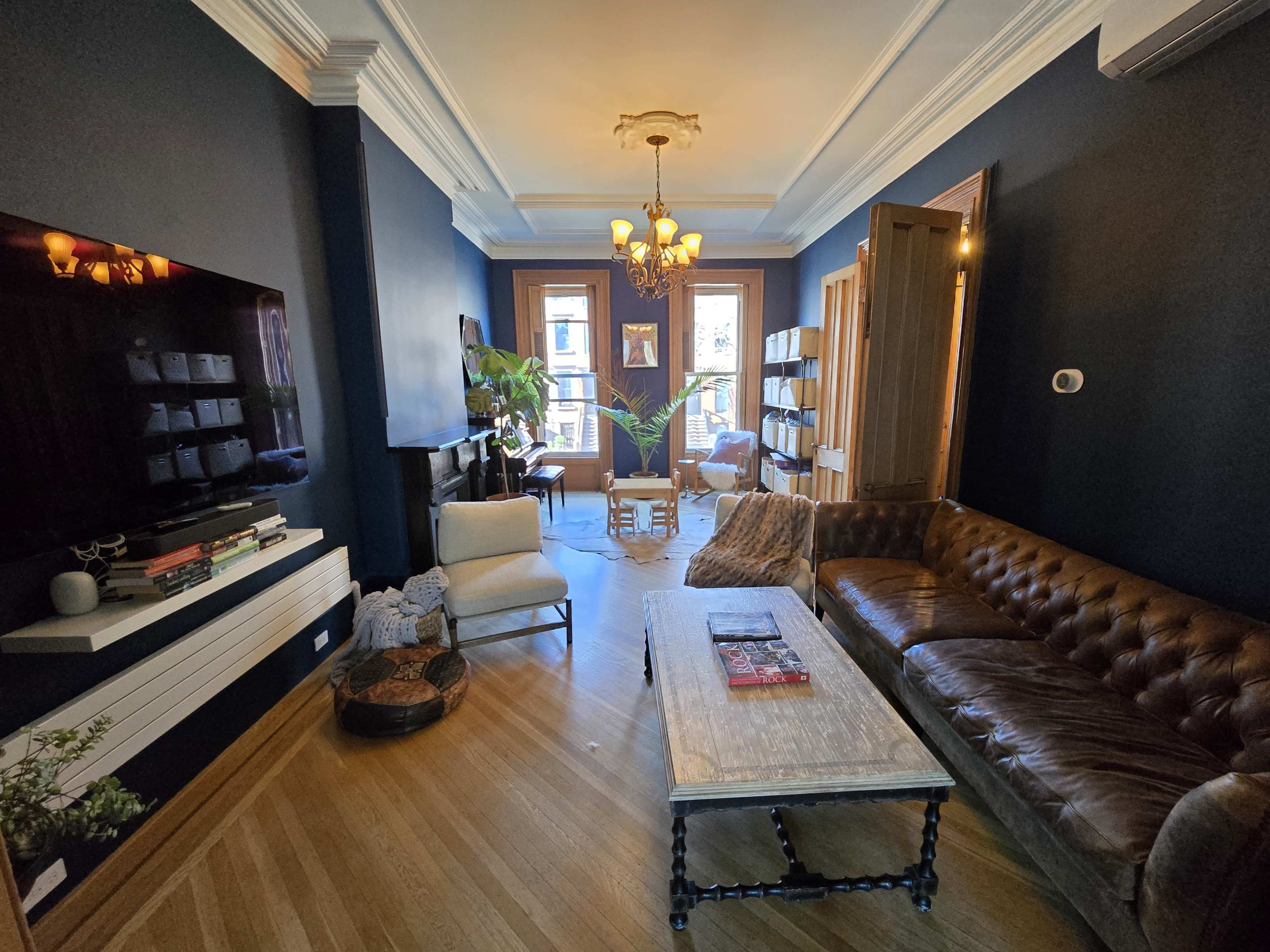 The image shows a stylish living room with a brown leather sofa, a wooden coffee table, and large windows that invite natural light, complemented by decorative plants and a chandelier.