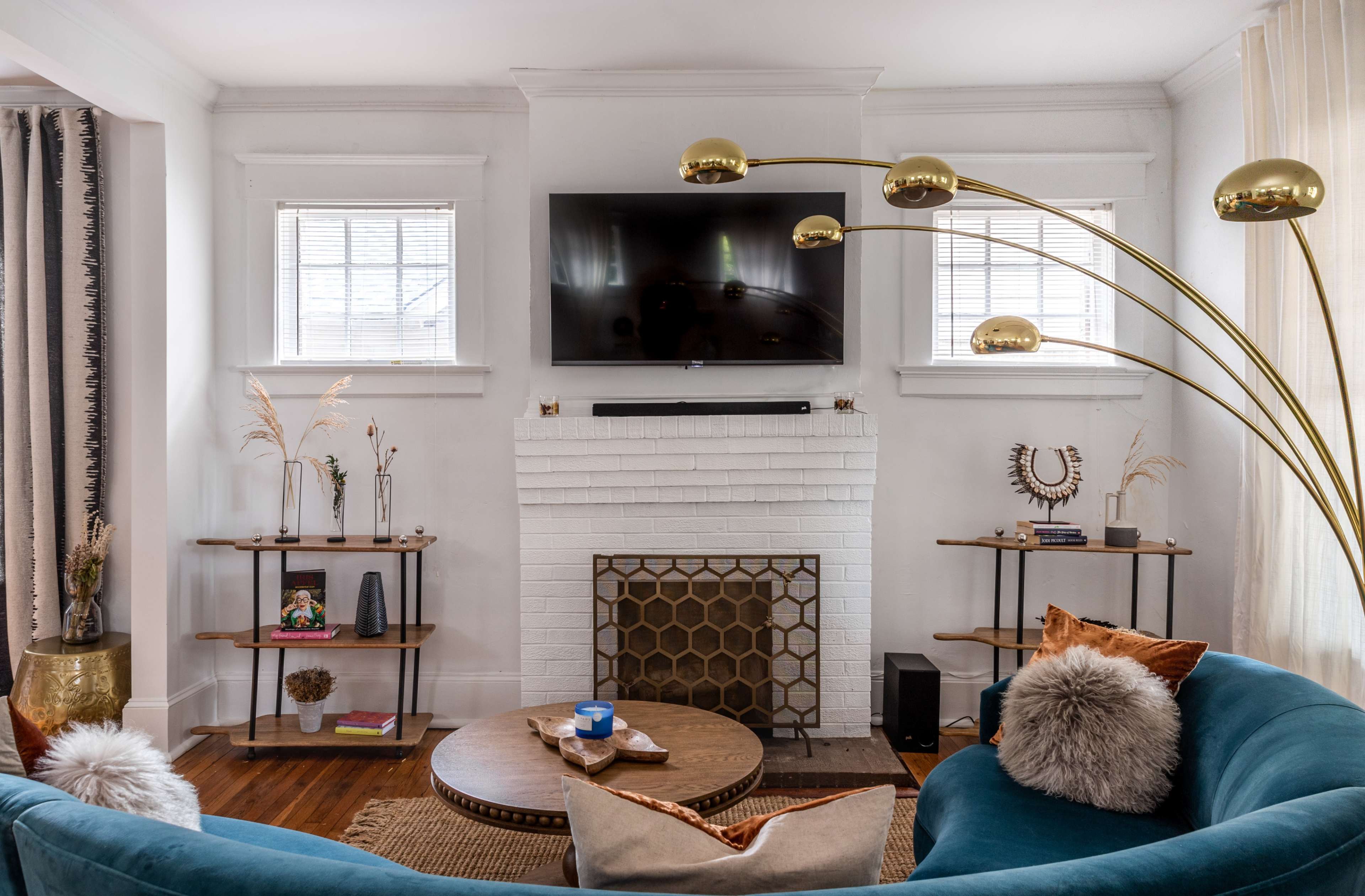 A modern living room features a teal sofa, a round coffee table, a gold-toned floor lamp, and a white-brick fireplace with a mounted television above it.