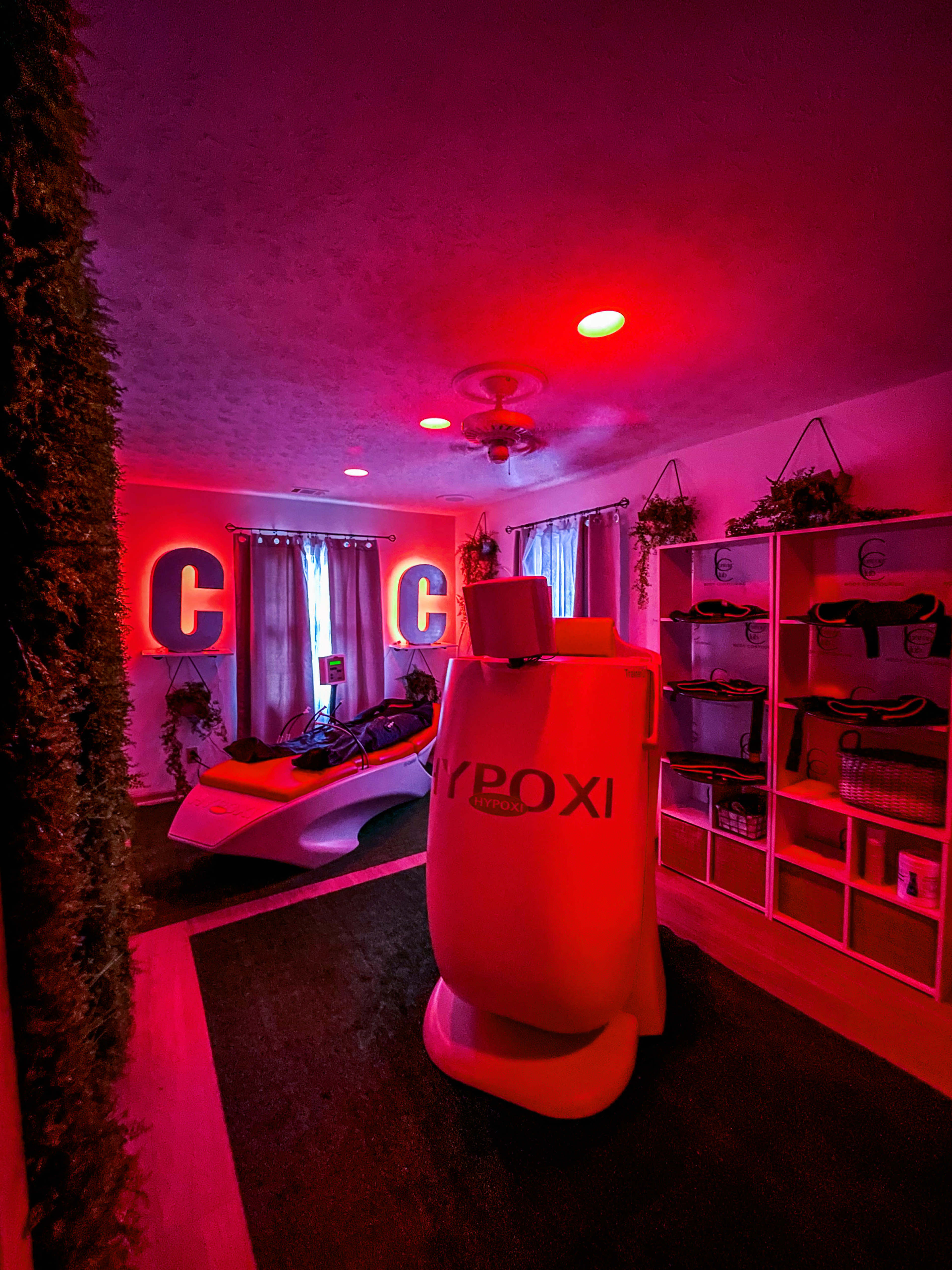 A dimly lit room featuring a white HYPOXI machine, shelves with fitness equipment, and large illuminated letters spelling "C C."