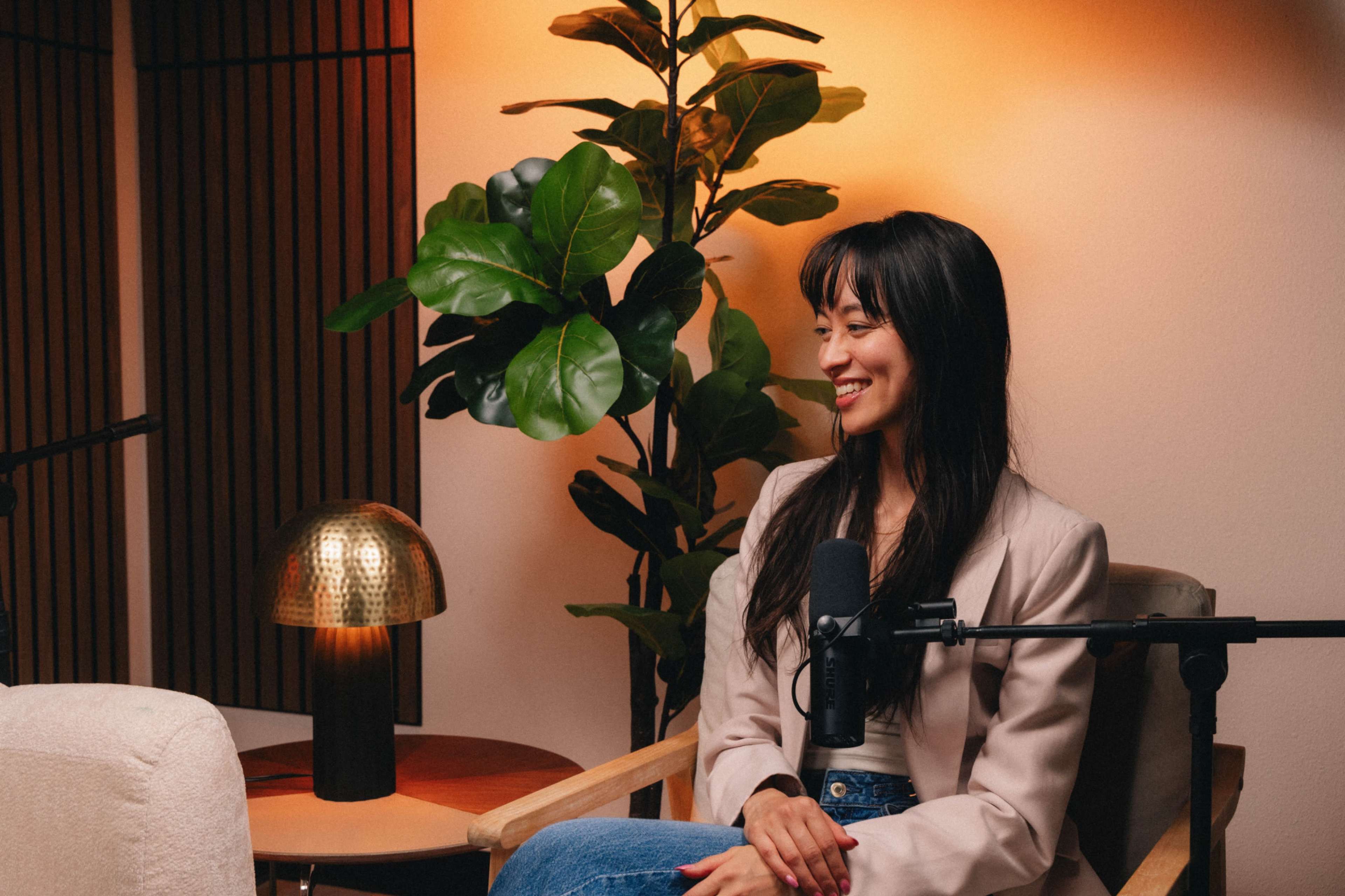 A woman sits in a chair beside a plant, with a microphone in front of her and warm lighting in the background.