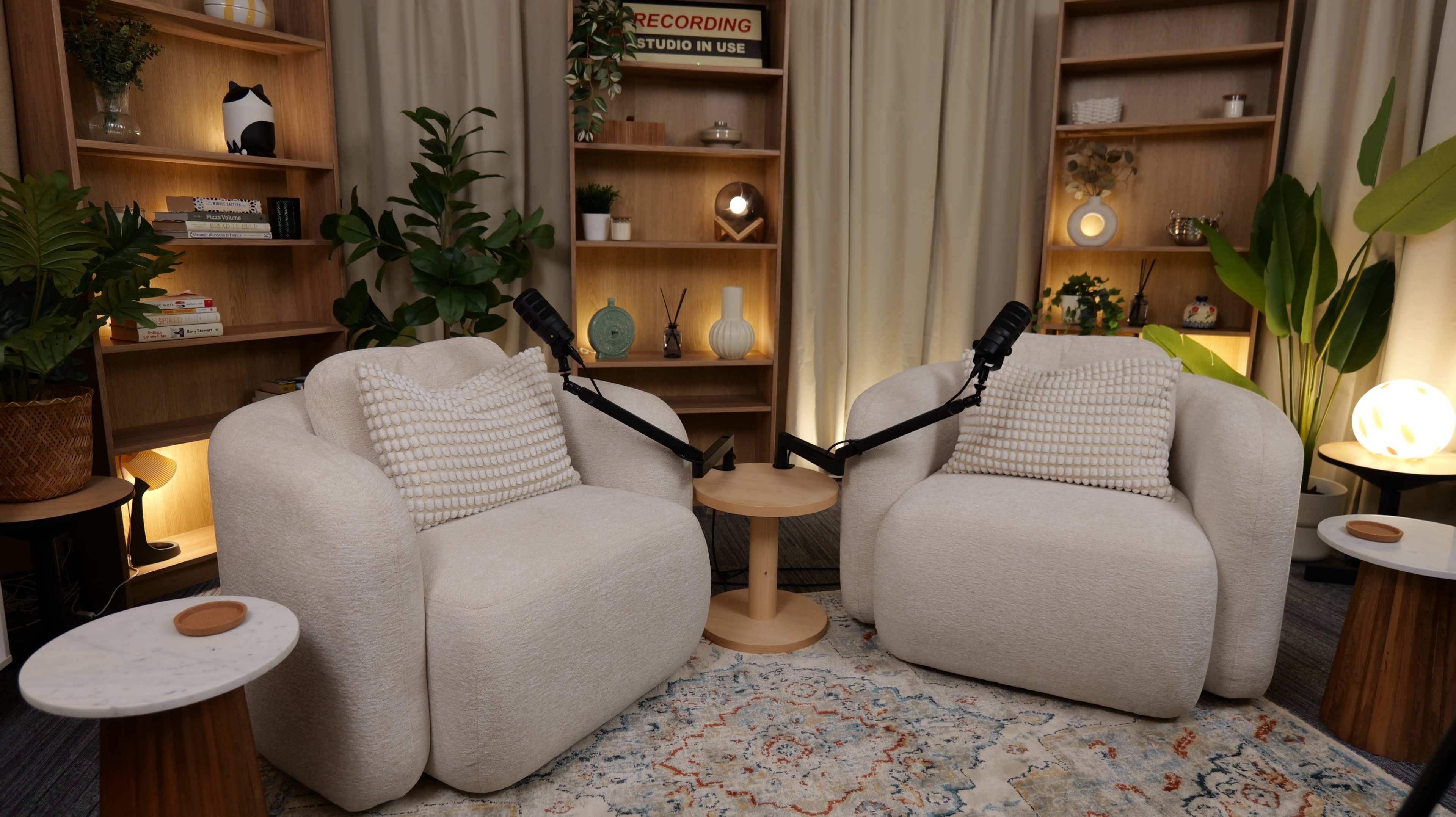The image depicts a cozy recording studio setup featuring two comfortable chairs with microphones positioned in front of them, alongside a small side table and decorative shelves in the background.