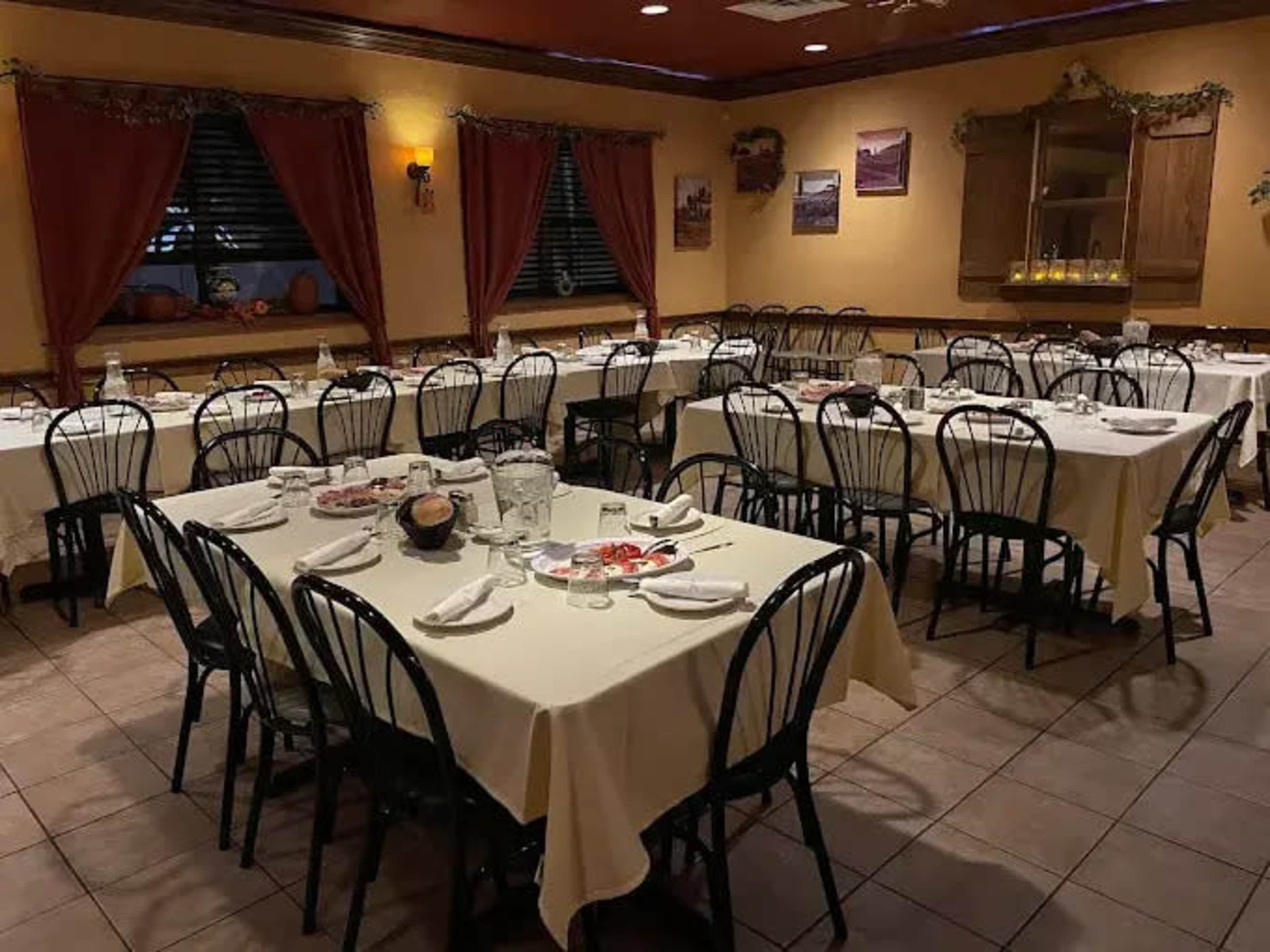 A dining room features multiple tables with white tablecloths set for a meal, surrounded by chairs and decorated with curtains and wall art.