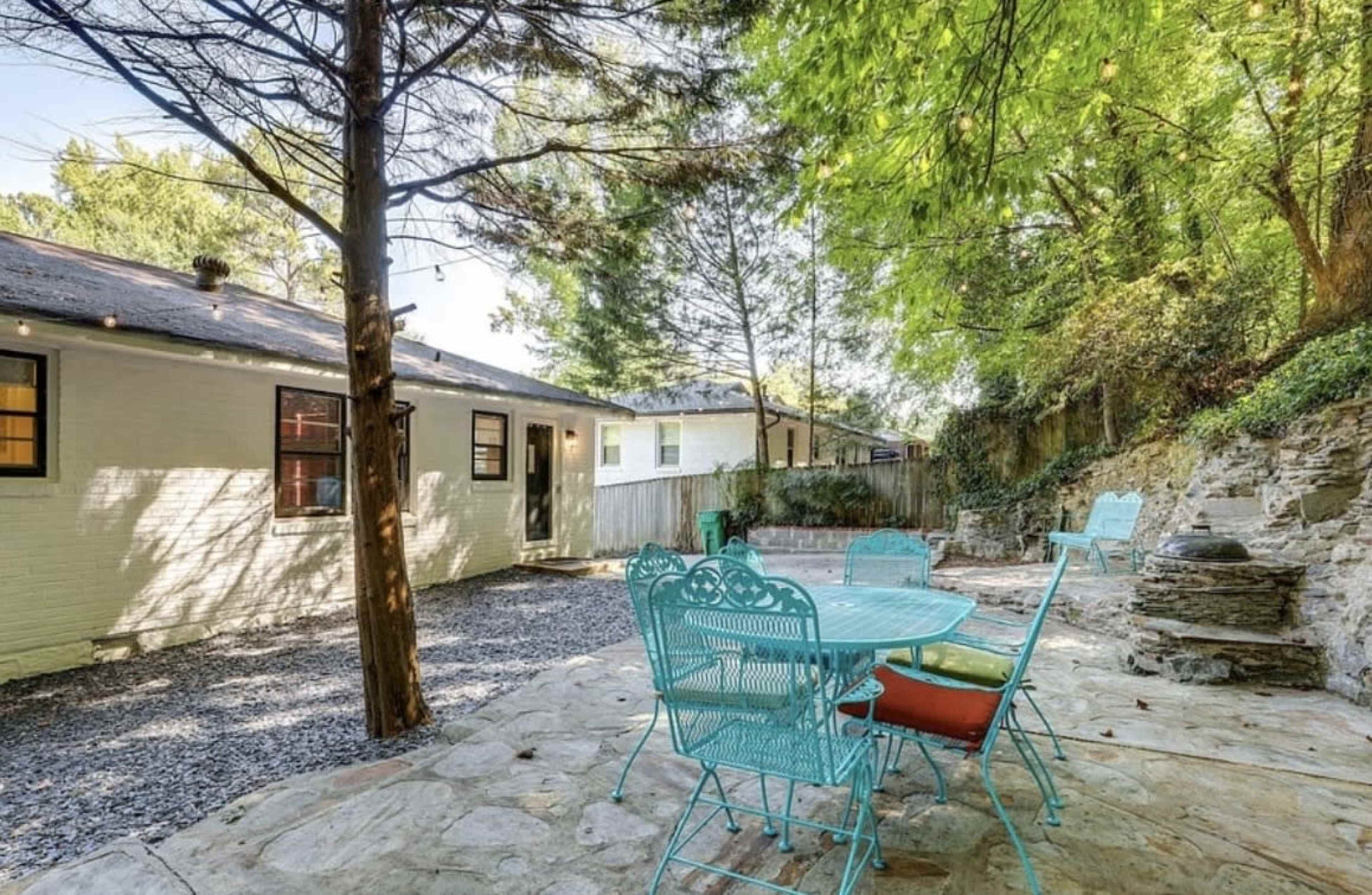 The image shows a stone patio with a round table surrounded by turquoise chairs, set in a garden area with trees and a stone wall in the background.