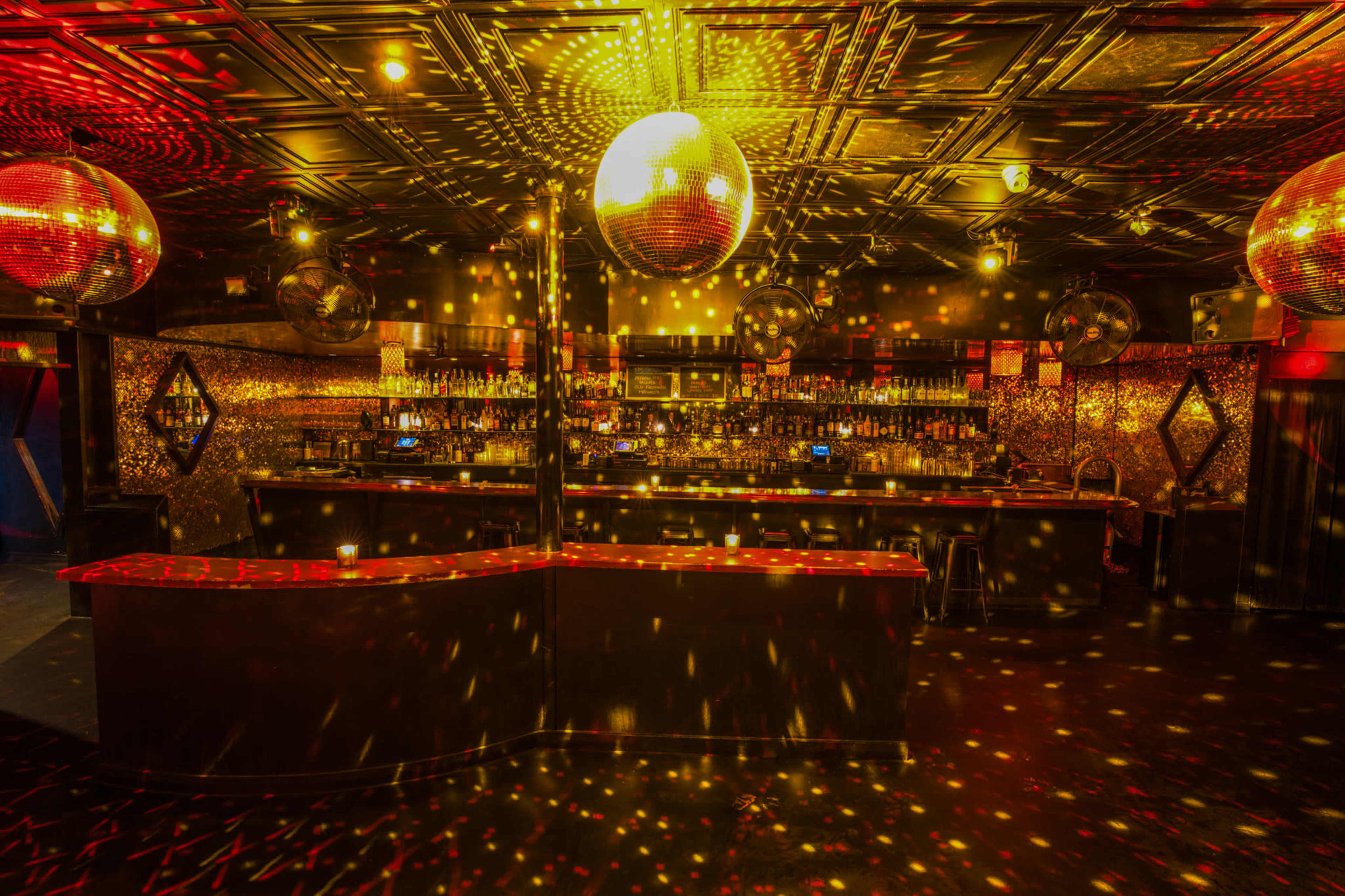 A dimly lit bar with a long counter, adorned with disco balls that cast colorful lights across the walls and floor.