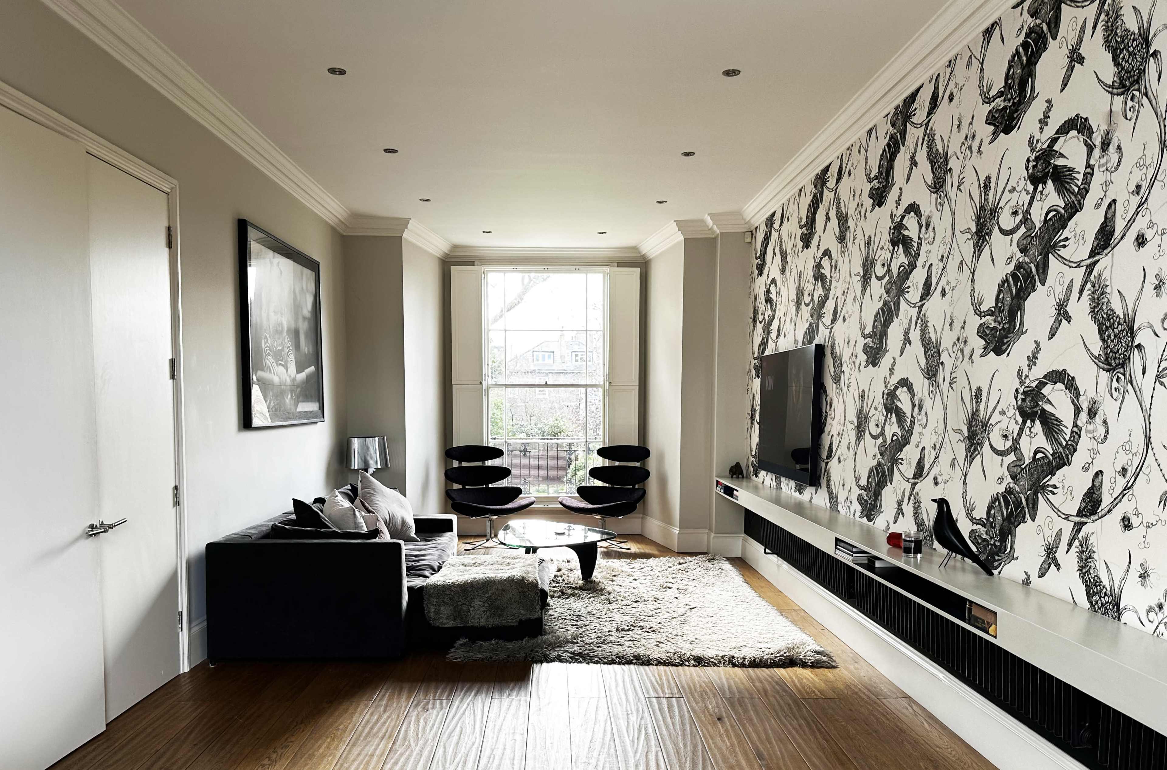 A modern living room with a large window, a black sofa, two black lounge chairs, a television mounted on a wall, and a textured wall featuring a floral pattern.